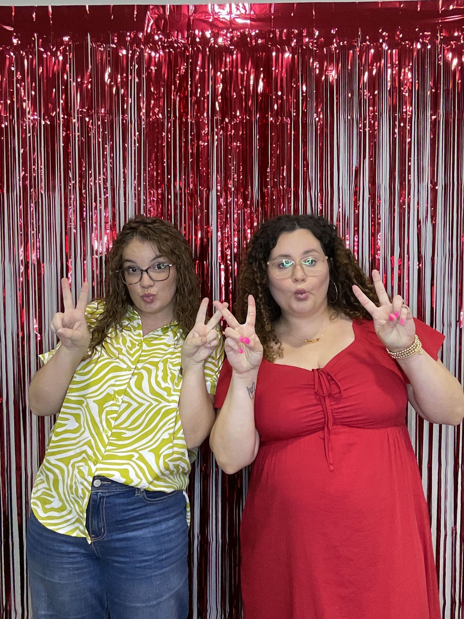 Two curly-haired women wearing glasses pose with peace signs in front of a red metallic fringe backdrop.