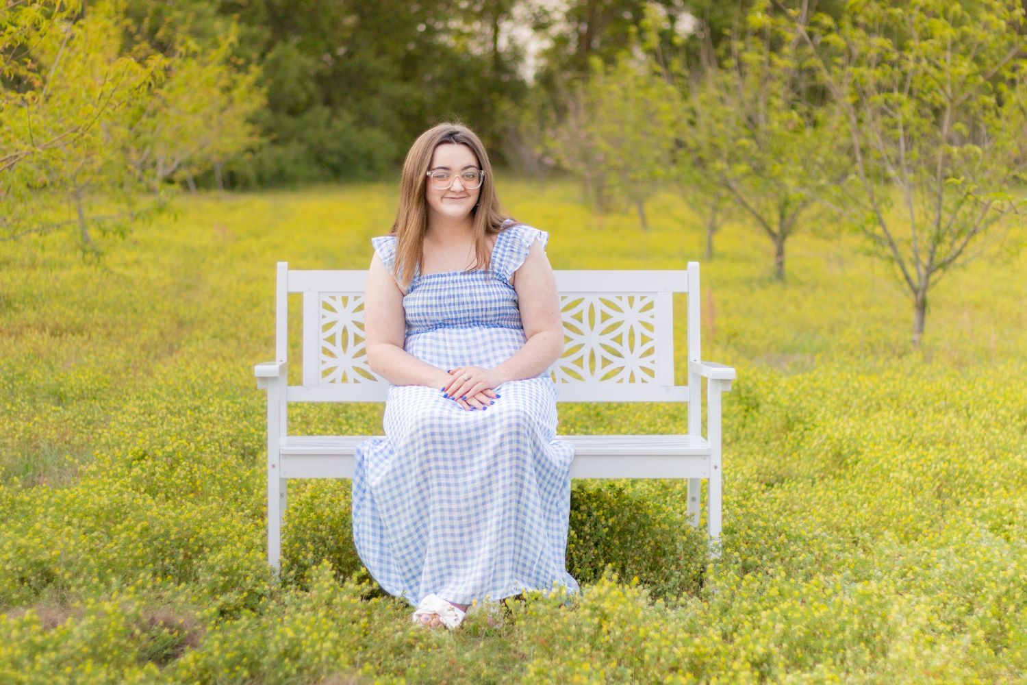 Smiling young woman in blue gingham maxi dress sitting on white decorative bench in yellow meadow.