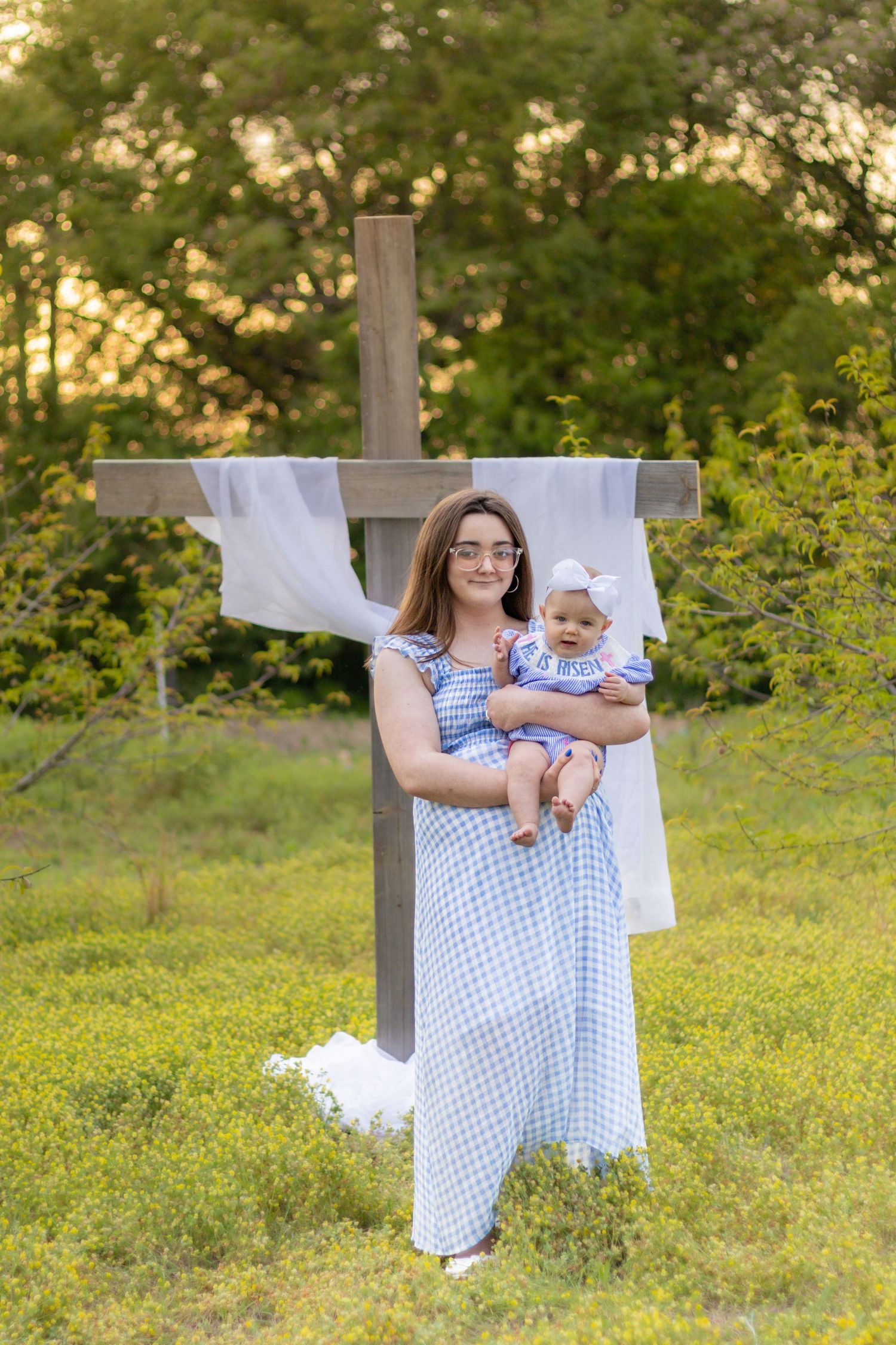 Smiling mother in blue gingham dress holding baby girl in matching outfit in front of a wooden cross outdoors.
