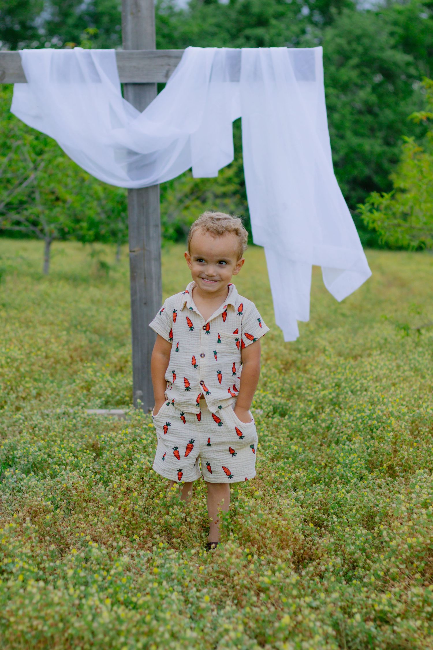 Cheerful young boy in carrot-print outfit stands smiling in a green meadow beneath white fabric draped on a wooden post.