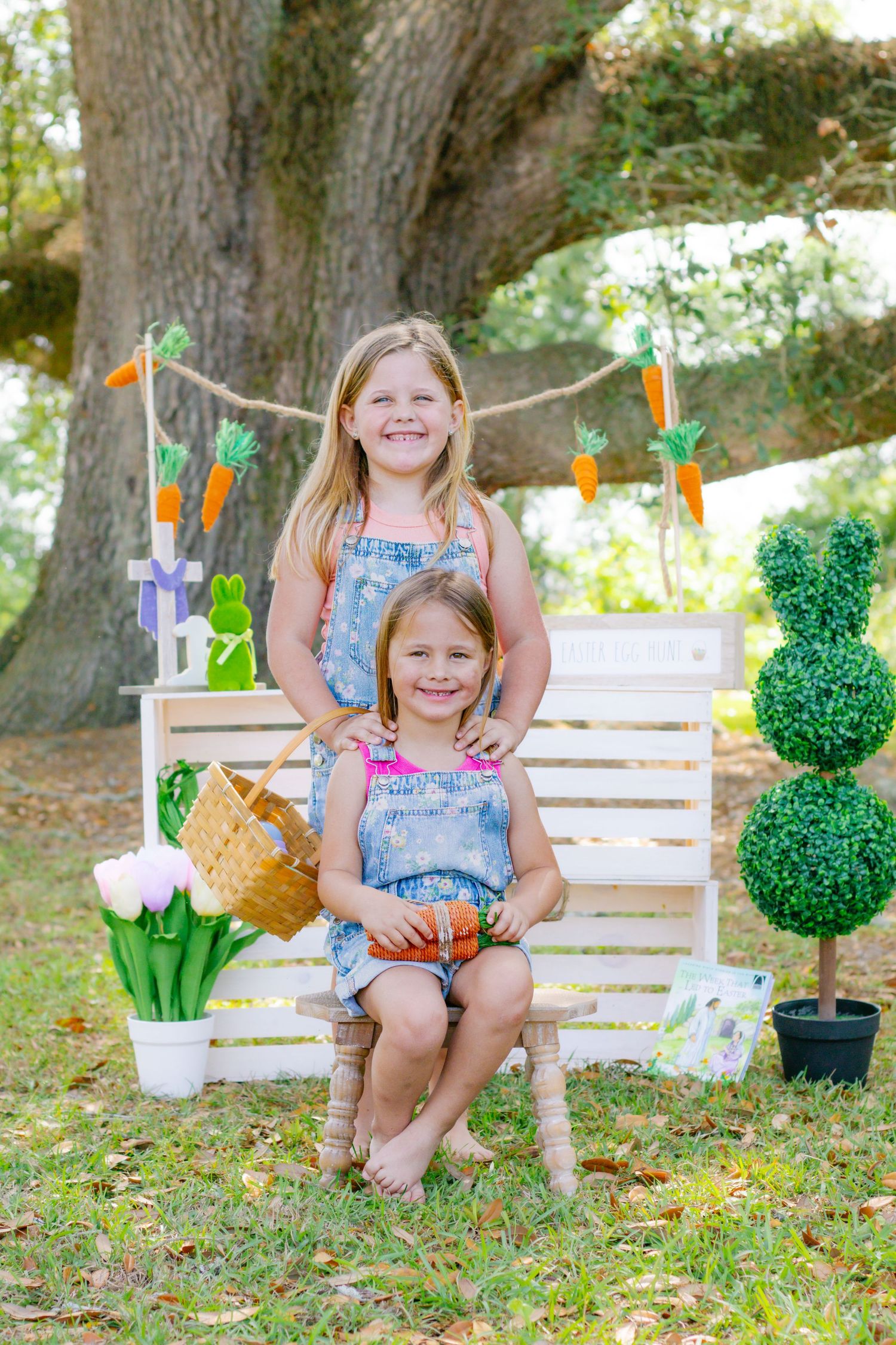 Two sisters in denim overalls pose at Easter photo set with carrot garland and bunny topiary.