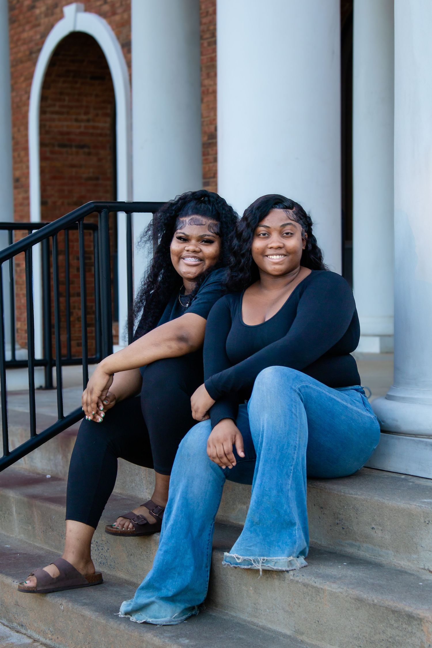 Two young Black women in black tops and jeans smile while sitting on brick building steps.