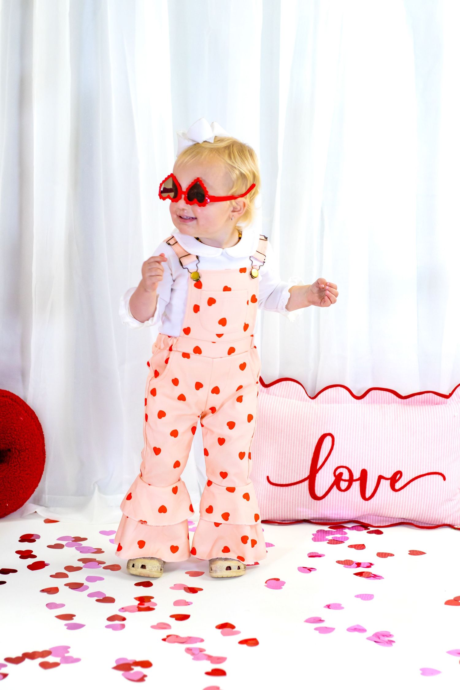Toddler girl in pink heart overalls and red heart sunglasses standing among Valentine's Day confetti.