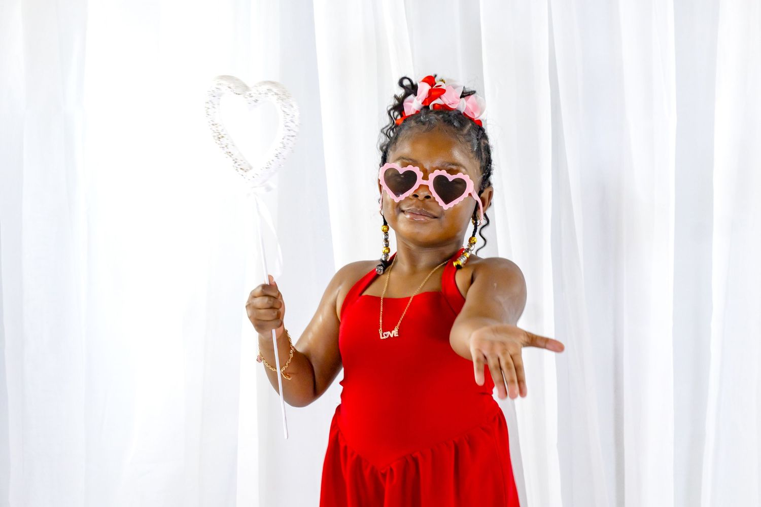 Young Black girl in red dress posing with heart-shaped sunglasses and wand prop.