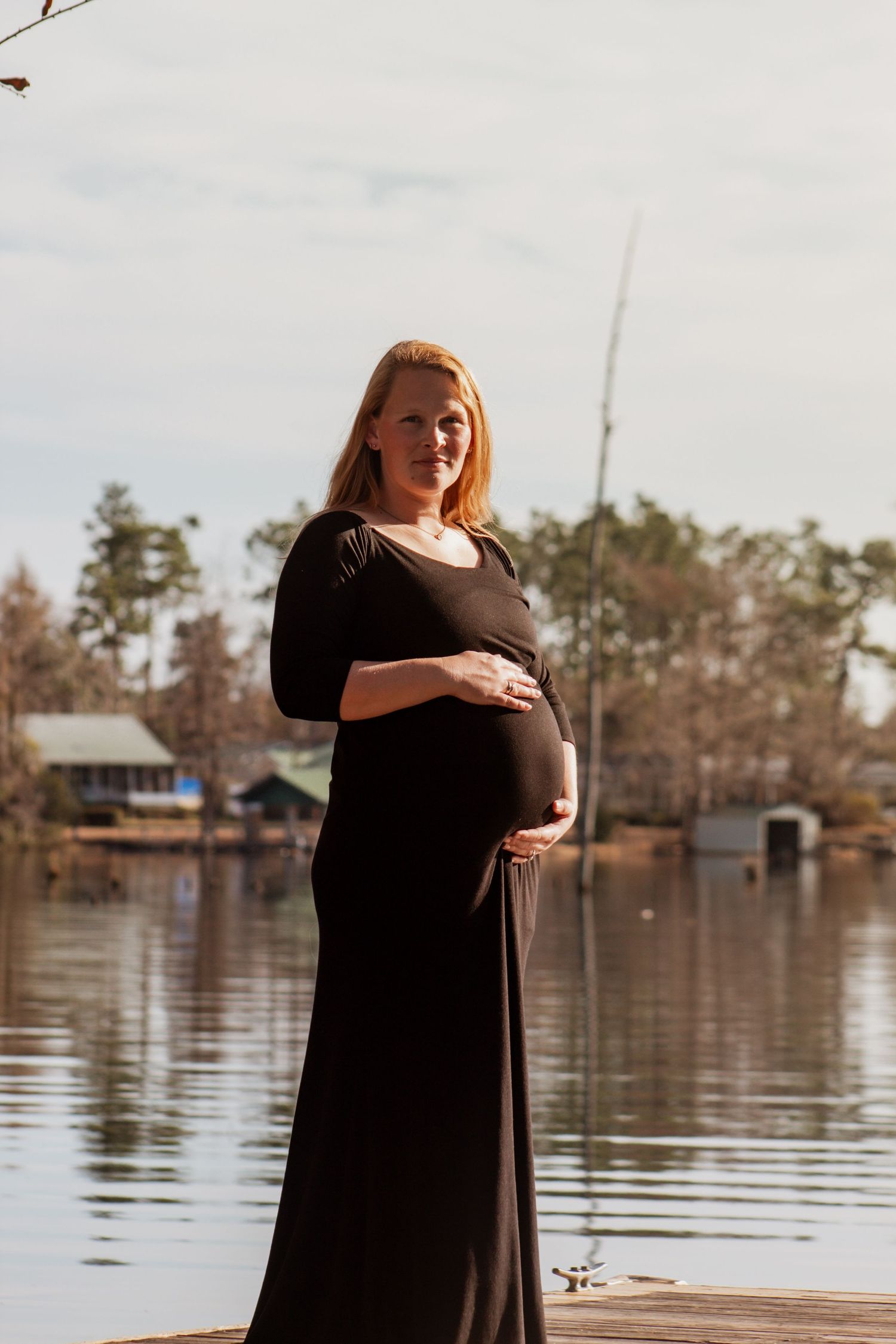 Smiling pregnant woman in black dress cradling baby bump on wooden dock with lake and trees in background.