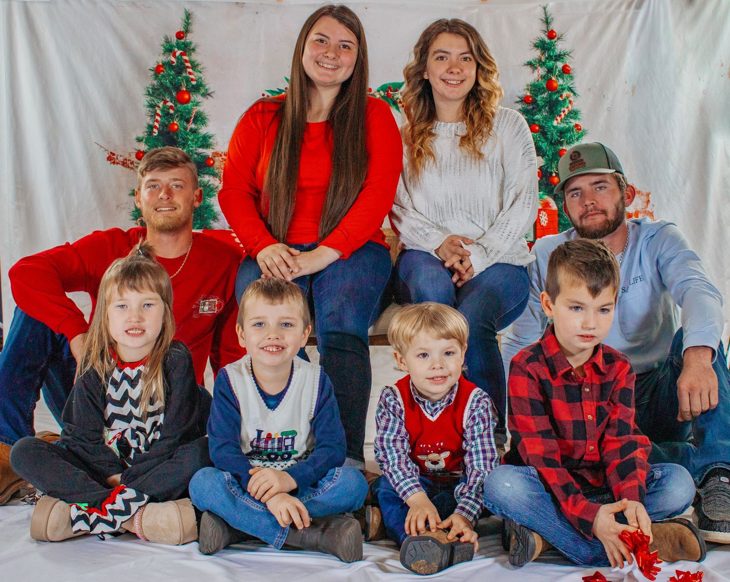 A cheerful family of eight poses together in front of Christmas trees for a holiday portrait.
