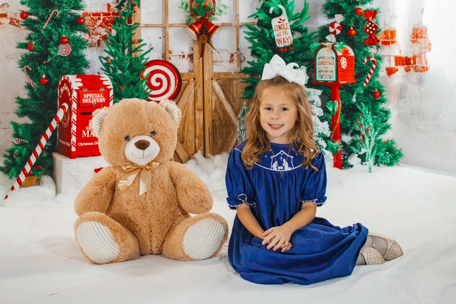 Smiling girl in blue velvet dress sitting beside large teddy bear in a festive Christmas photo studio.