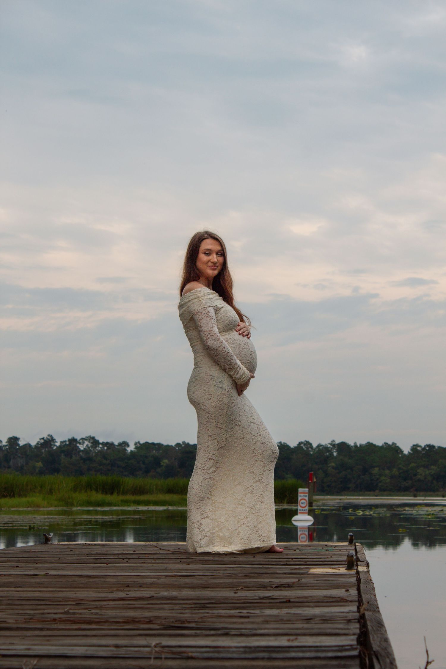 Smiling pregnant woman in white lace gown standing on wooden dock by lake, cradling her baby bump.