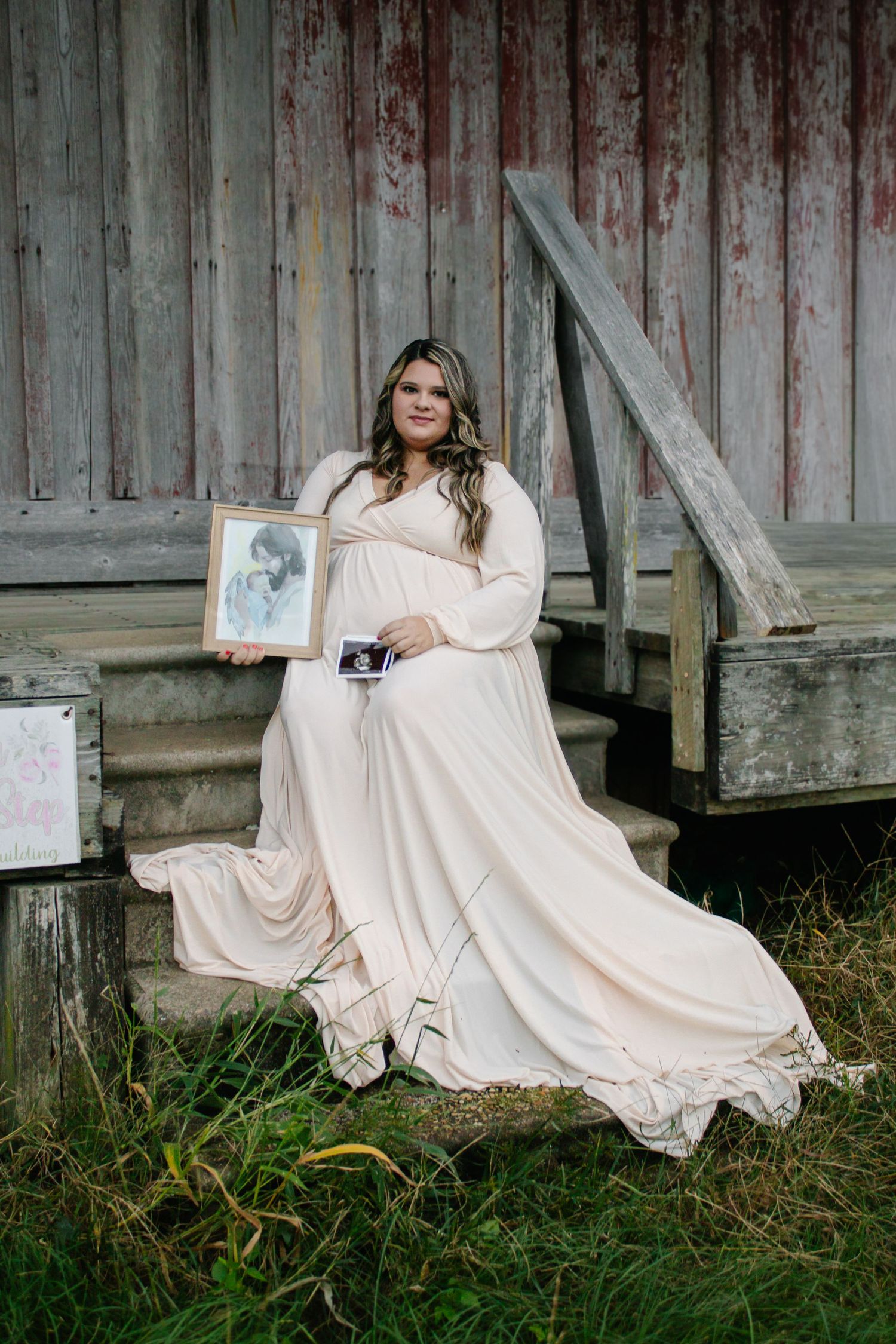 Pregnant woman in cream gown holding ultrasound and portrait on barn steps.