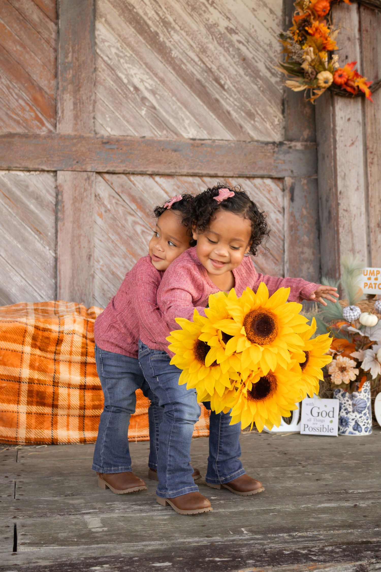 Twin girls in pink sweaters holding large sunflower bouquet in front of rustic barn backdrop.