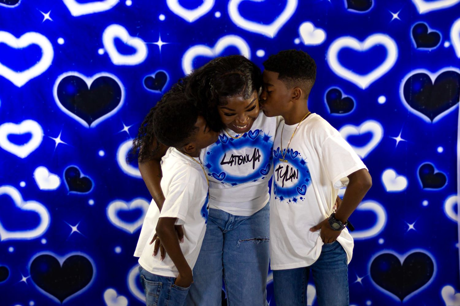 Woman hugging two children in matching white t-shirts against a blue heart background.