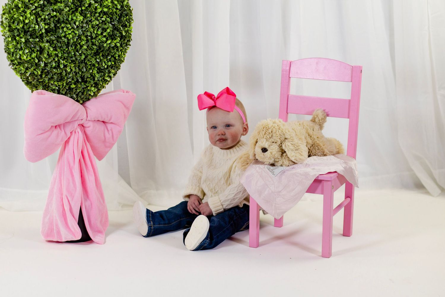 Baby girl in white sweater with pink bow sitting beside a pink chair with teddy bear and heart topiary.