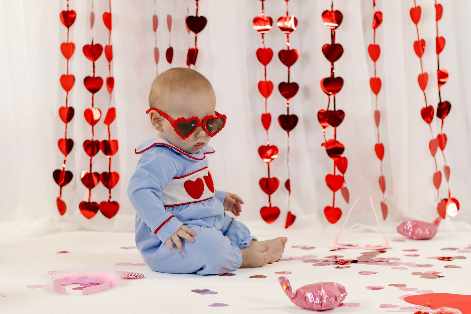 Baby in blue heart romper wears heart sunglasses while sitting among red hanging hearts and confetti.