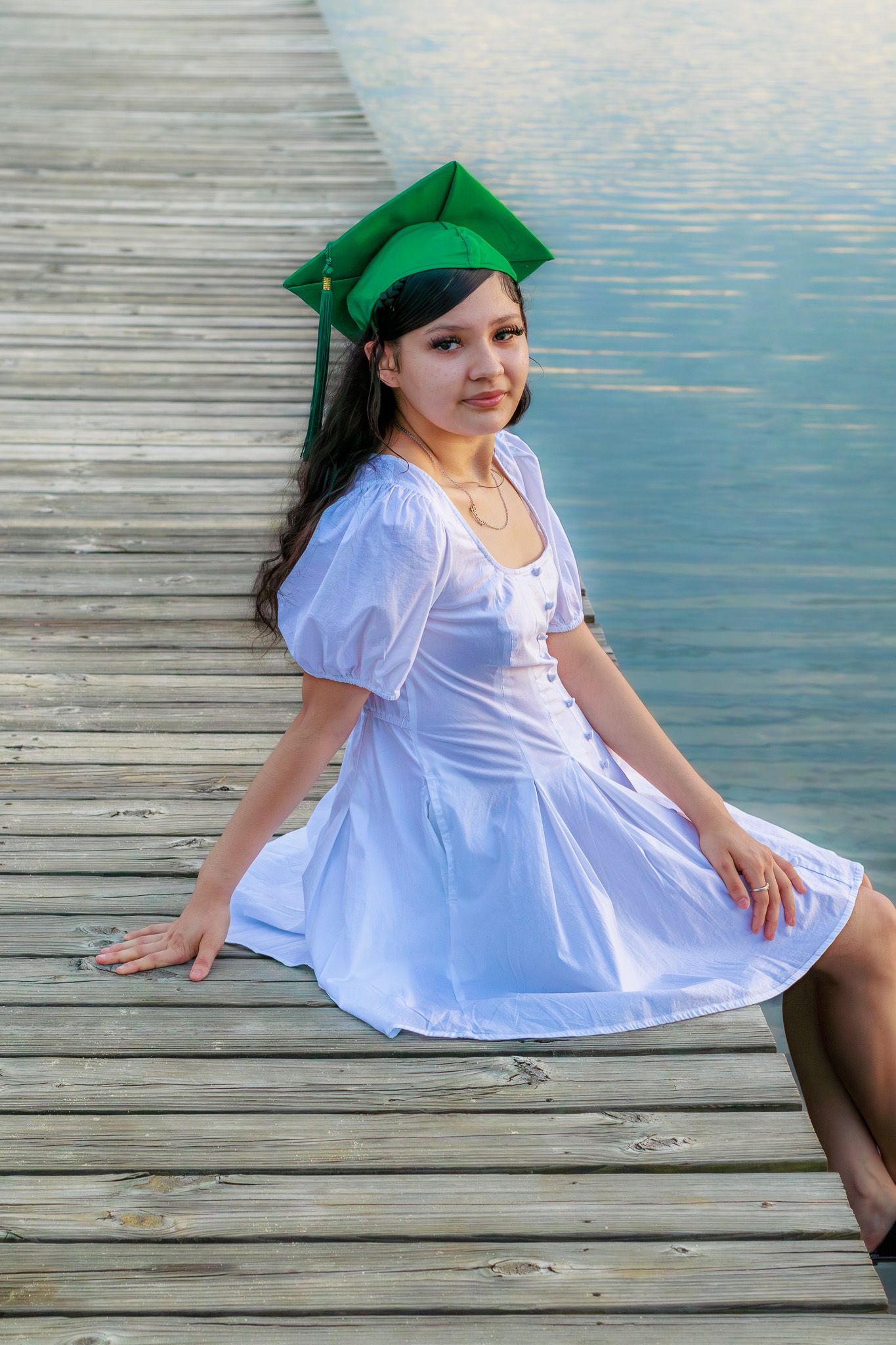Graduate in white dress and green cap sits gracefully on wooden pier beside calm lake waters.