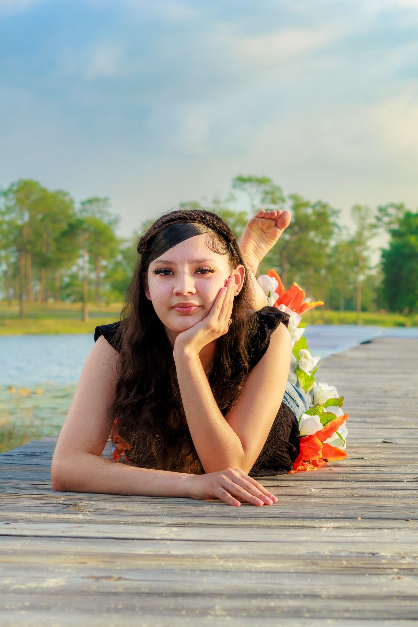 Young woman with dark hair lying on wooden dock by a lake, resting chin on hand, with flowers and trees behind her.