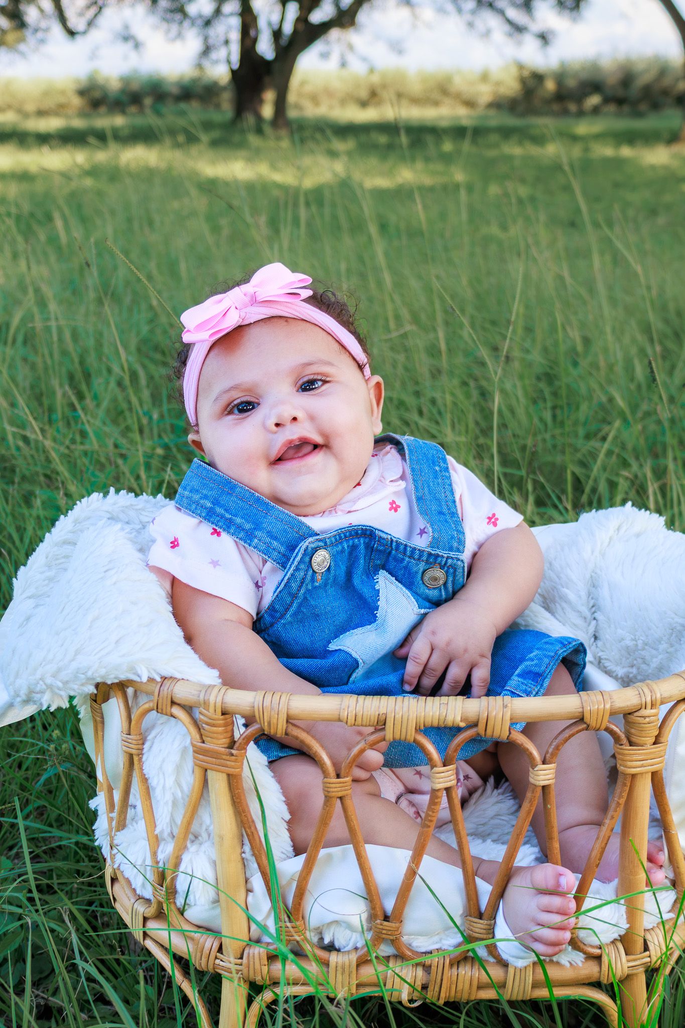 Adorable baby girl in denim overalls and pink bow headband sitting in a wicker basket outdoors.