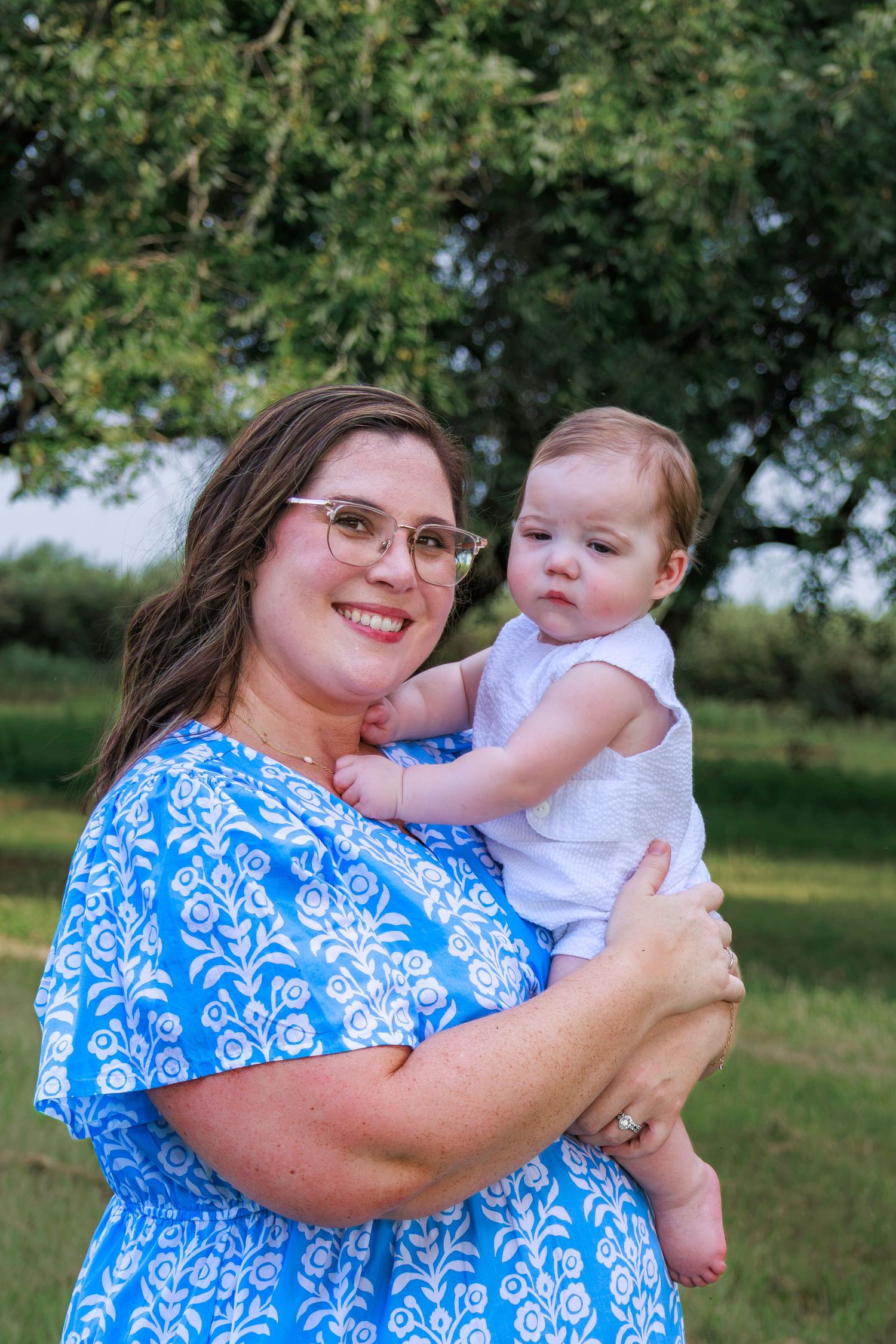 A smiling woman in a blue floral dress holds a baby in a white outfit outdoors near green trees.