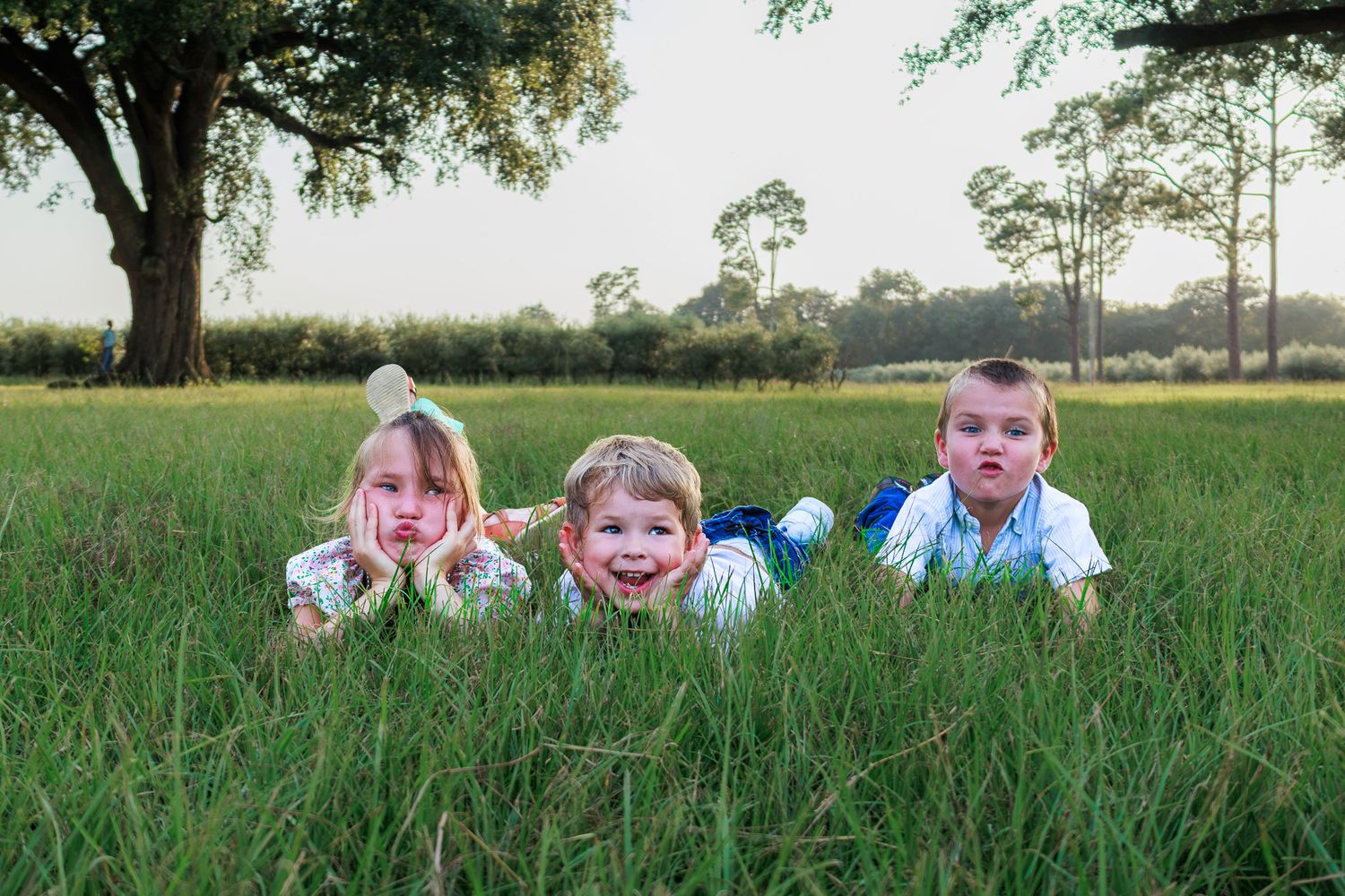 Three children lying in green grass field, smiling outdoors near large tree on sunny day.