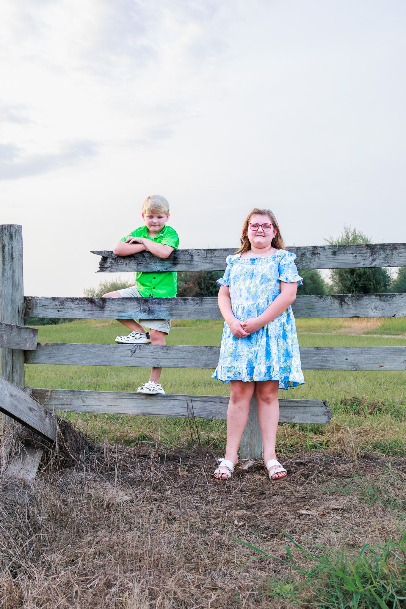 Two children pose by a wooden fence on a farm, a boy in green and a girl in a blue floral dress.