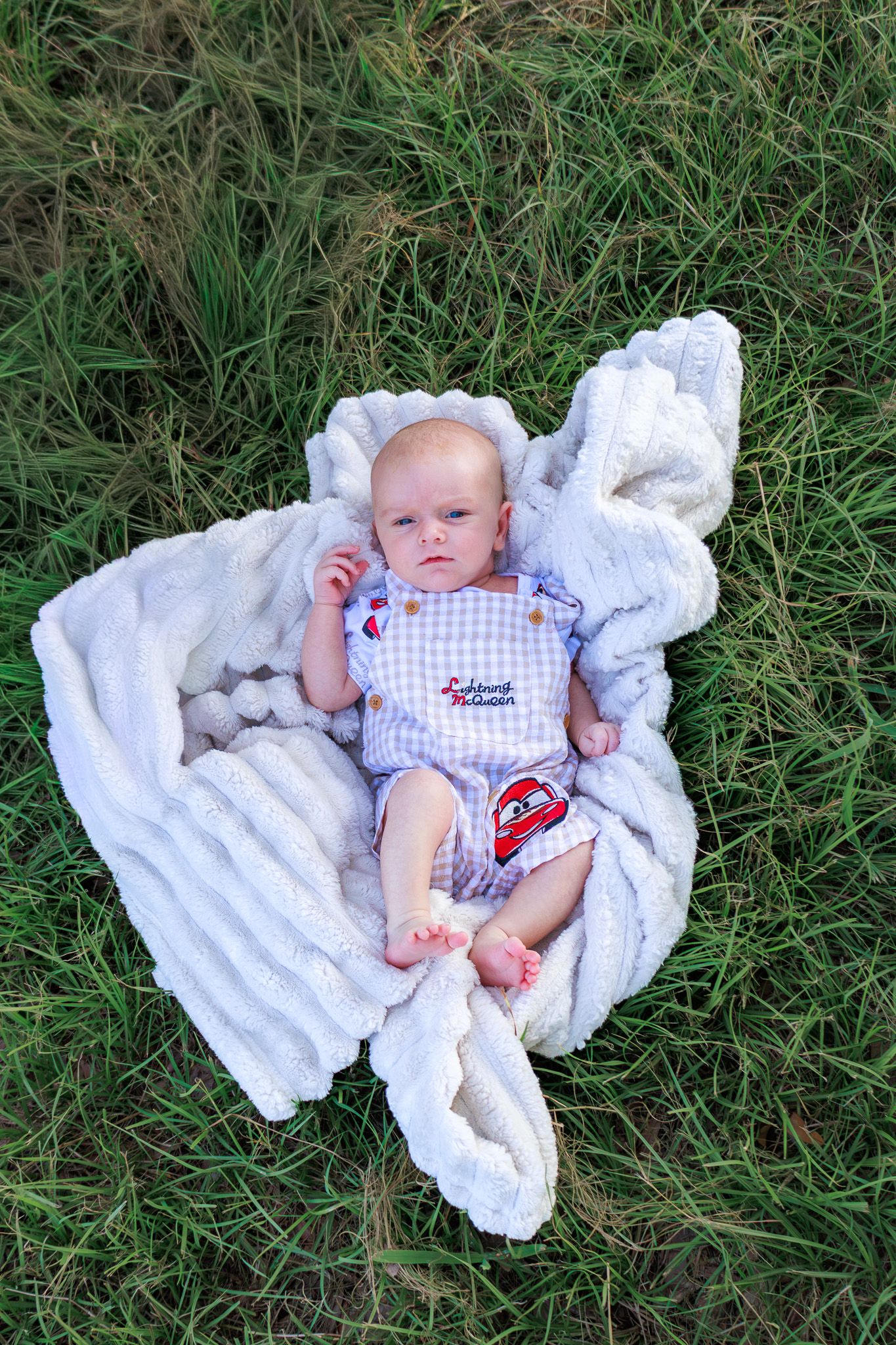 Newborn baby lies on white blanket in green grass wearing gingham outfit during outdoor photo session.