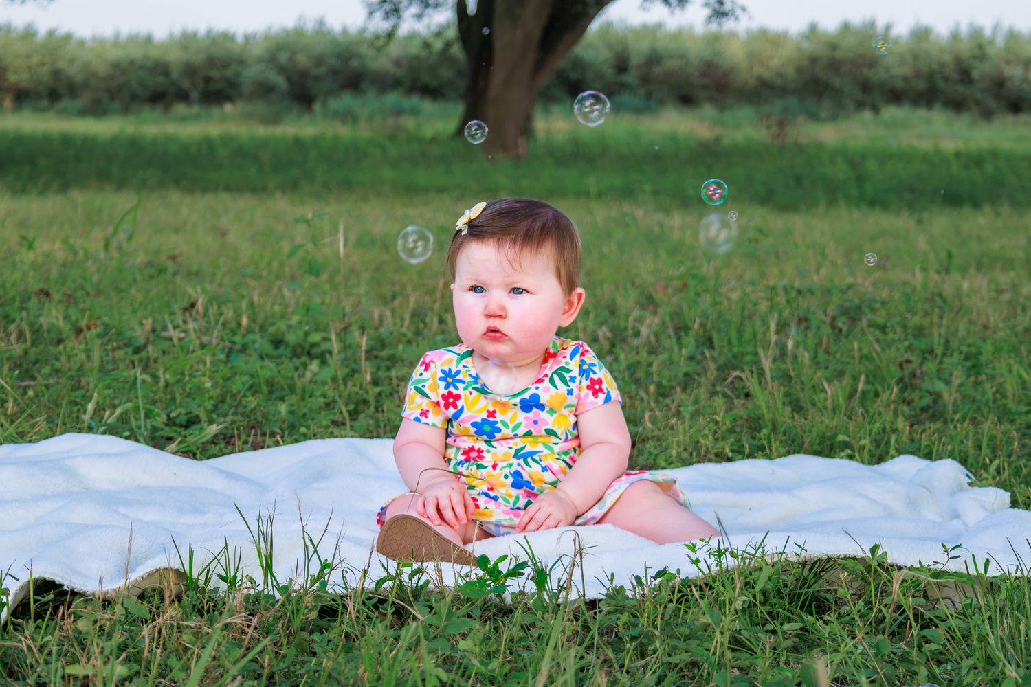 Cute baby girl sitting on white blanket in grass with bubbles floating around her.