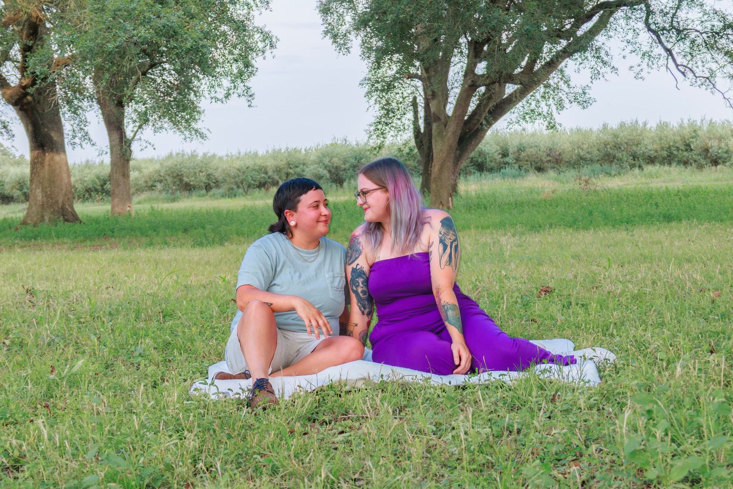 Two women sitting on a blanket in a green park, smiling at each other near trees on a sunny day.
