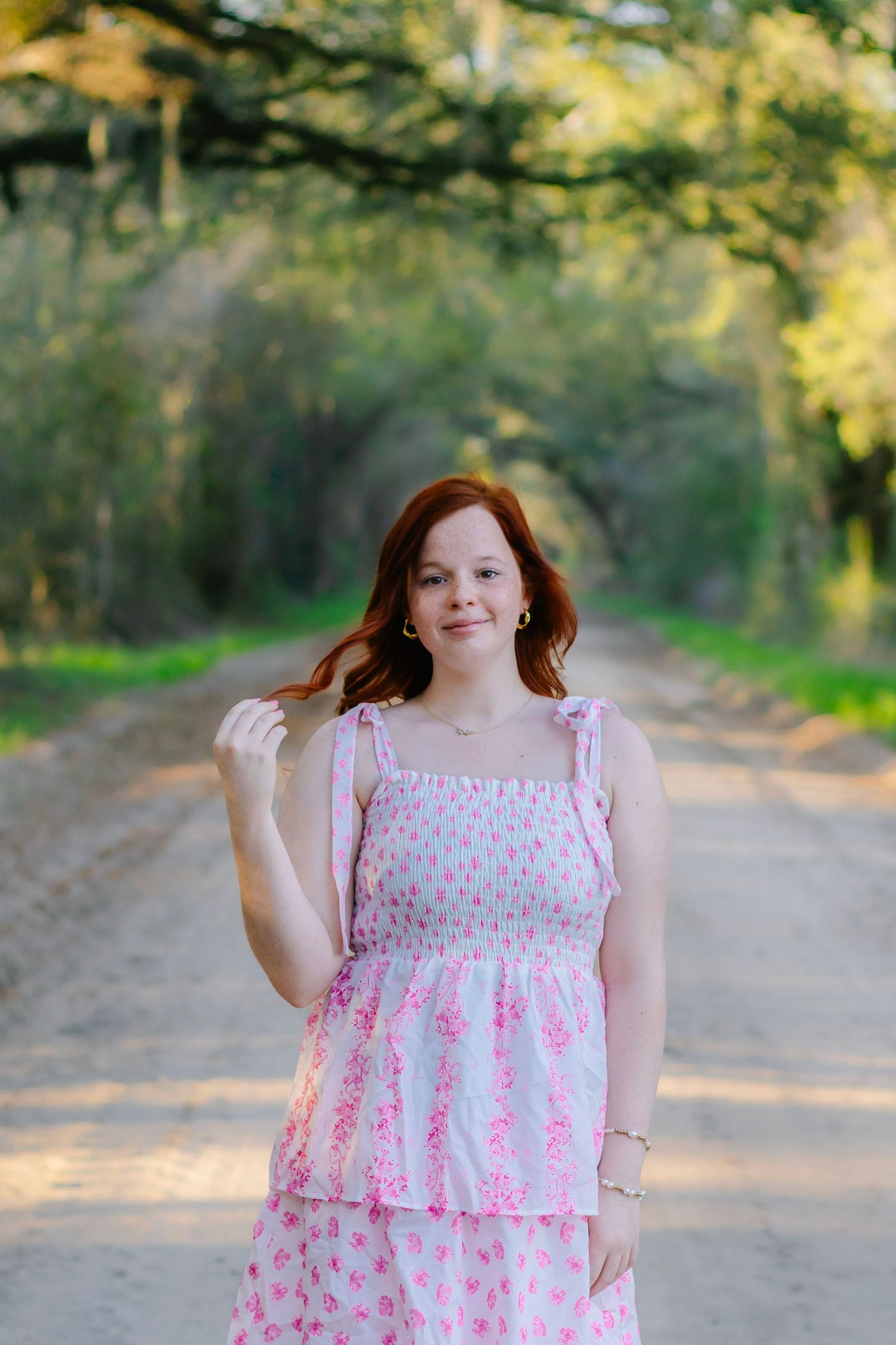 Smiling redhead girl in a pink floral dress stands on a sunlit tree-lined dirt road.