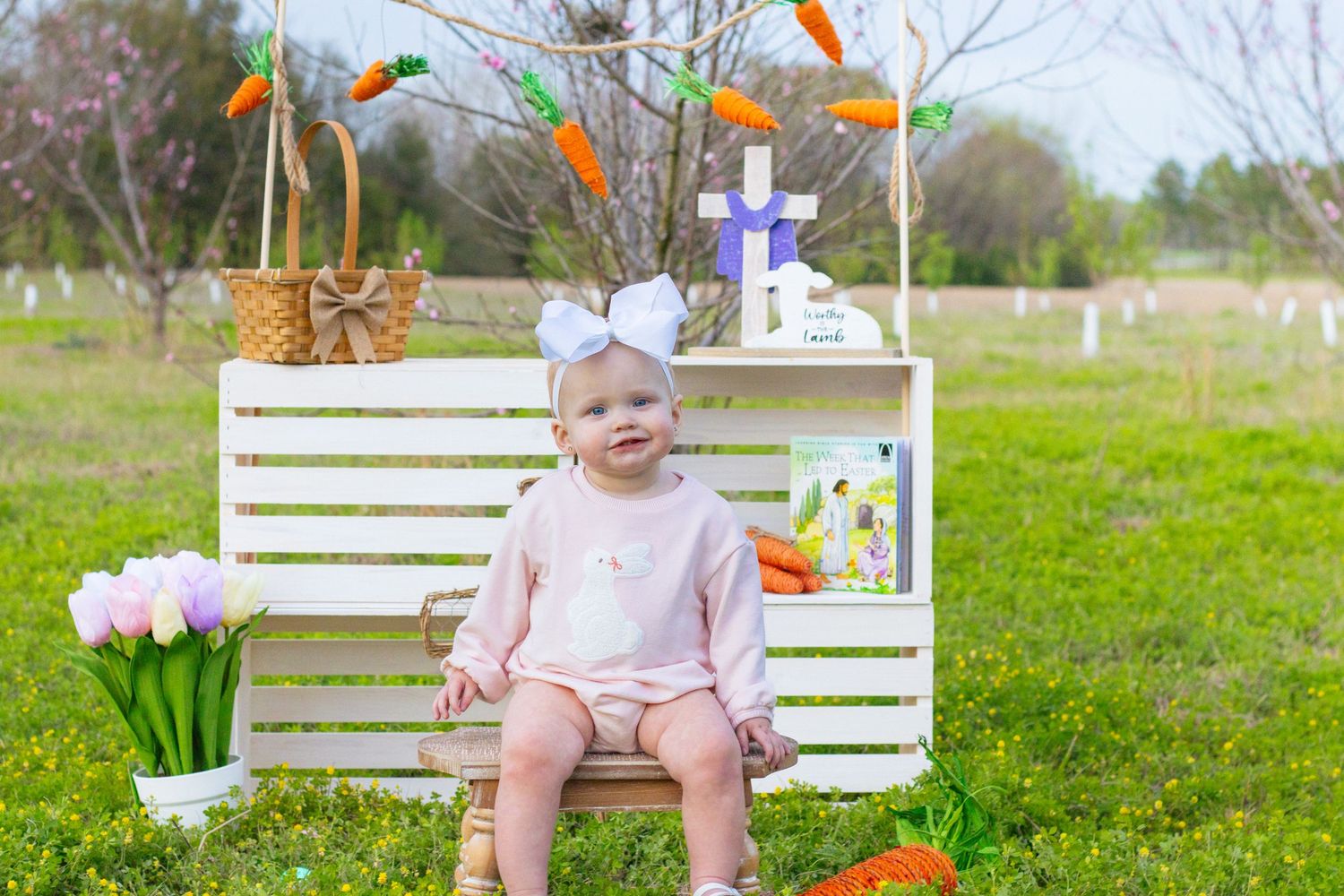 Baby with white bow headband sits on bench surrounded by Easter decorations in garden.