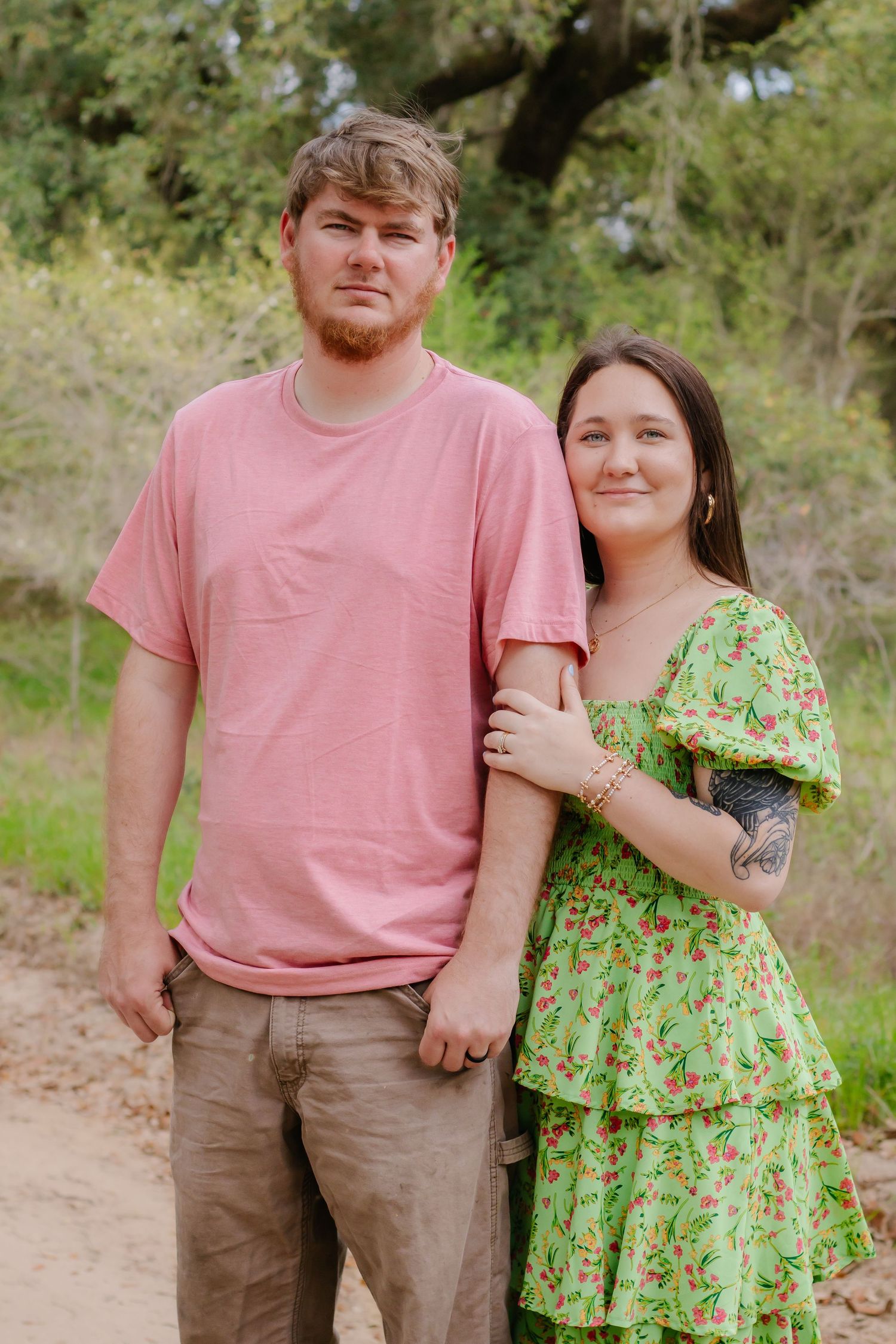 Young couple posing together on a nature trail, woman in green floral dress and man in pink shirt.