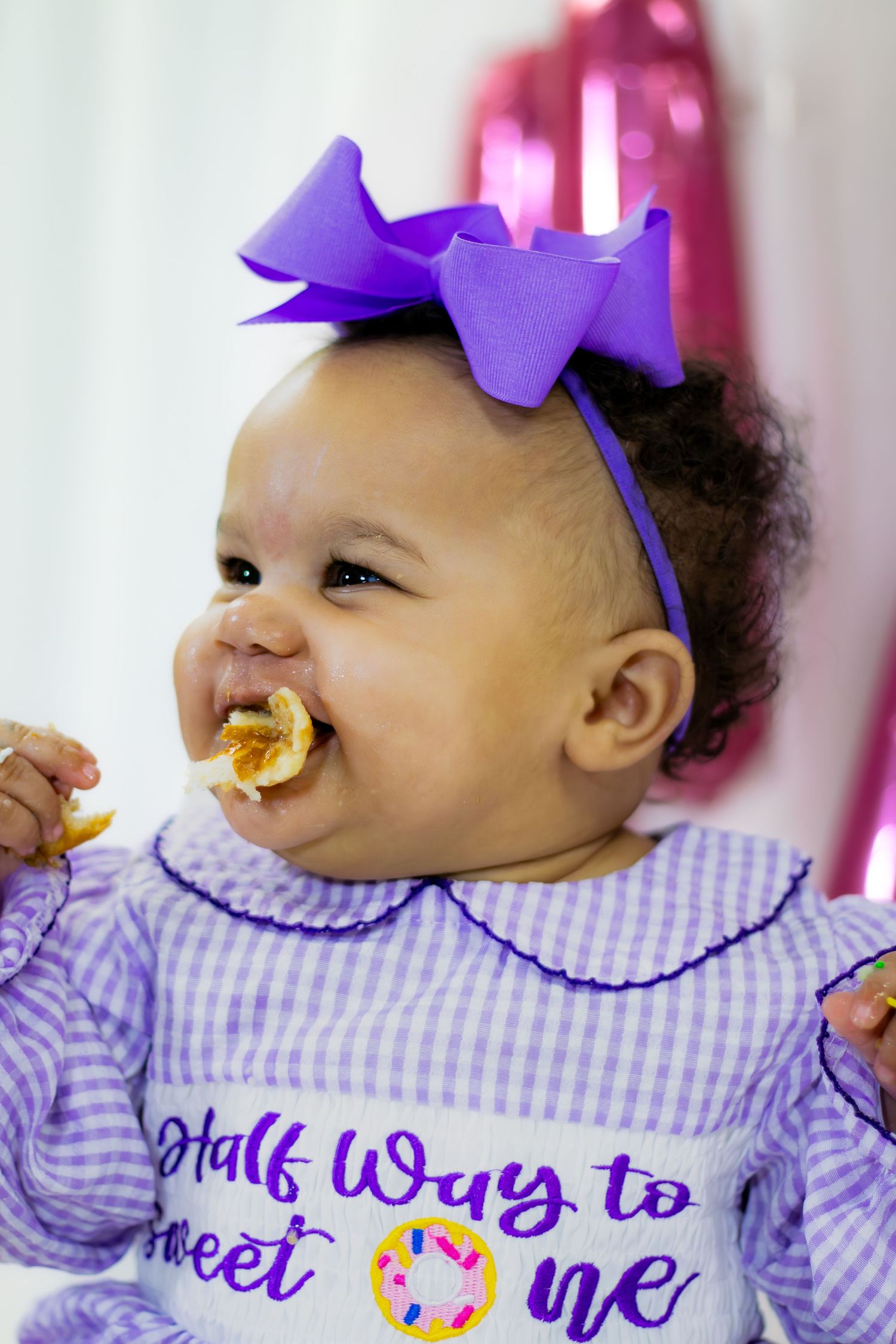 Baby girl with purple bow headband eating donut wearing Half Way to Sweet One romper.