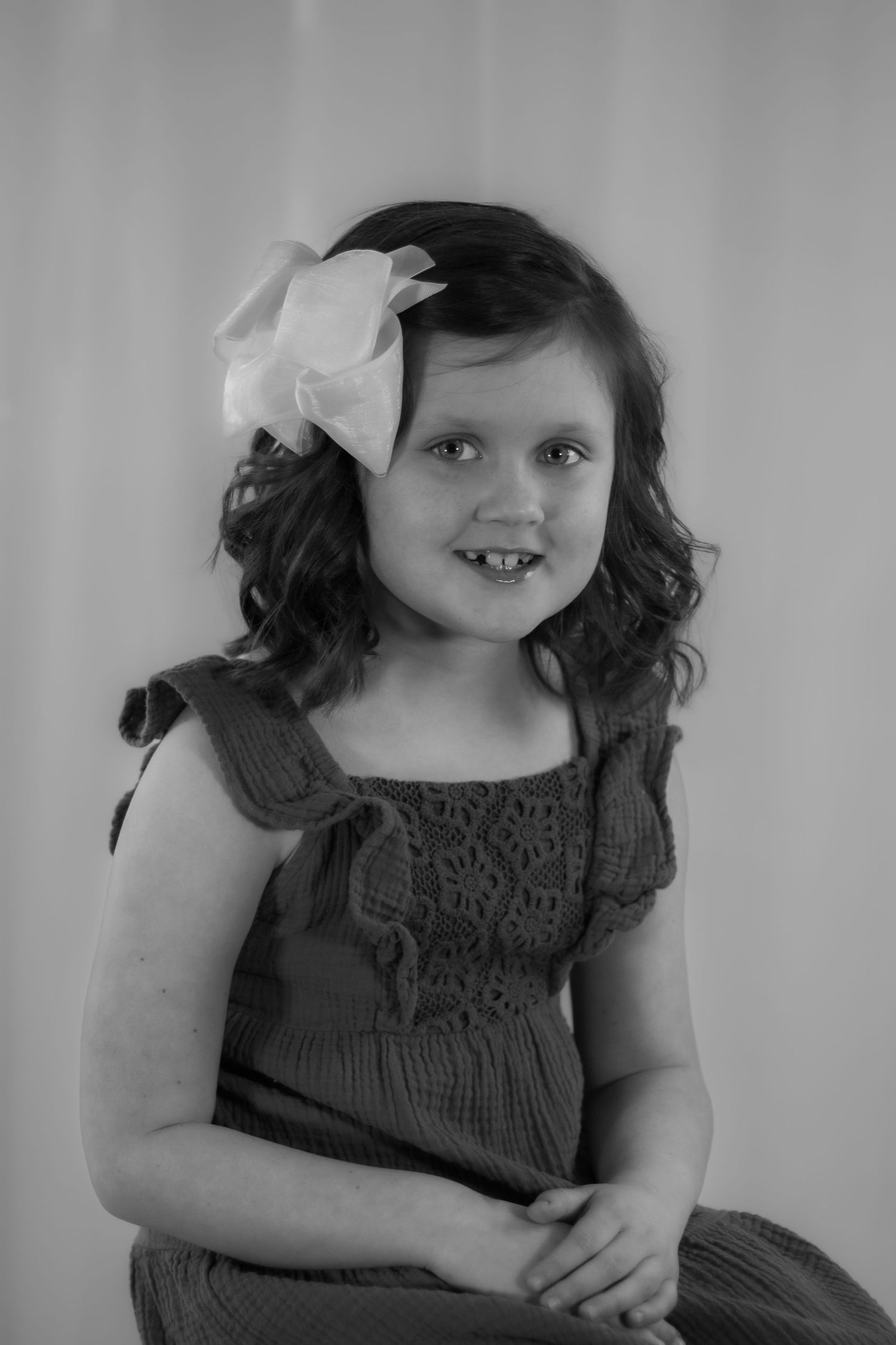 A sweet young girl with curly hair and a white bow smiles in a classic black and white portrait.