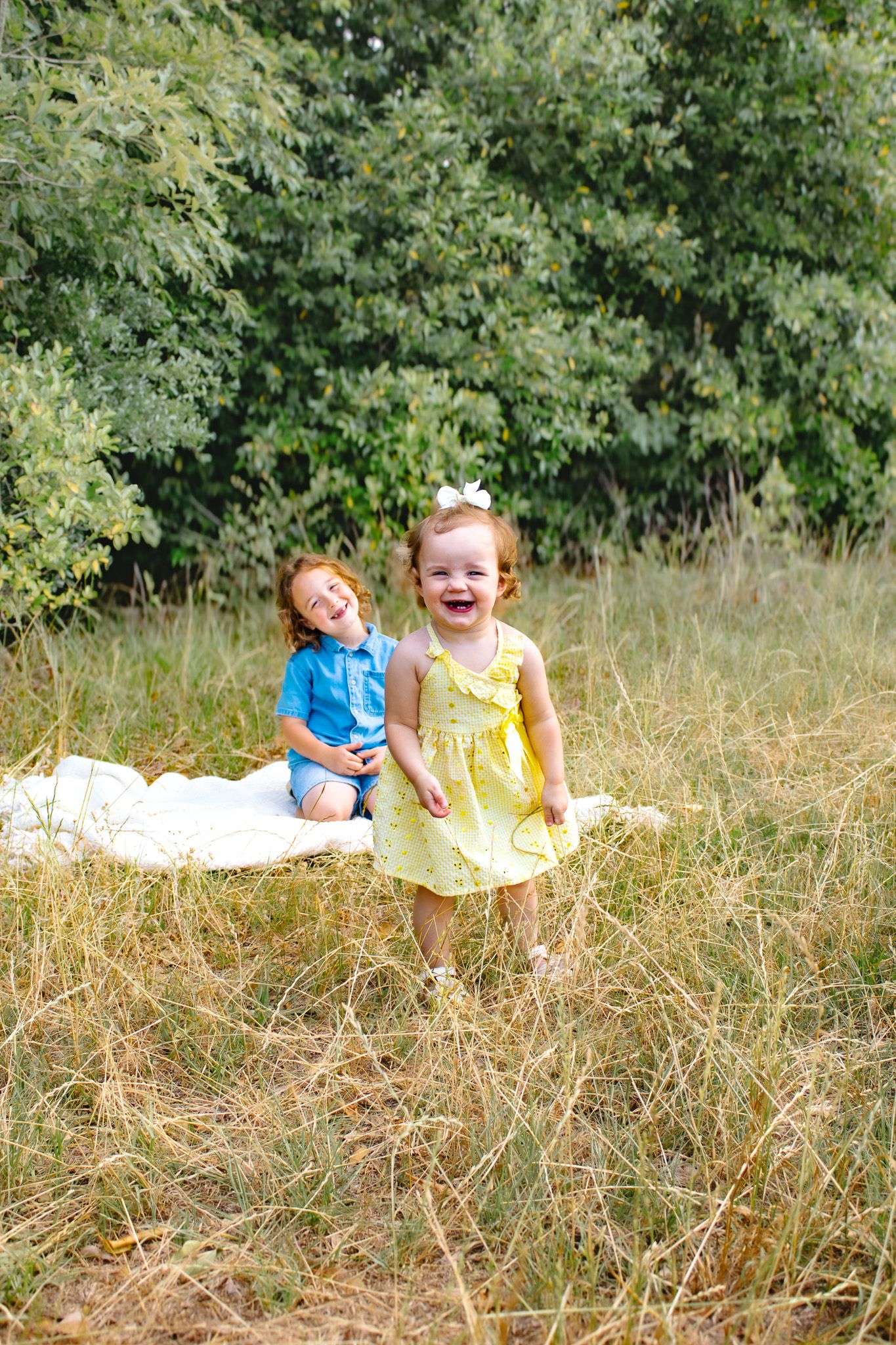 Two laughing redhead children play outdoors on a white blanket in a grassy field with green trees behind them.