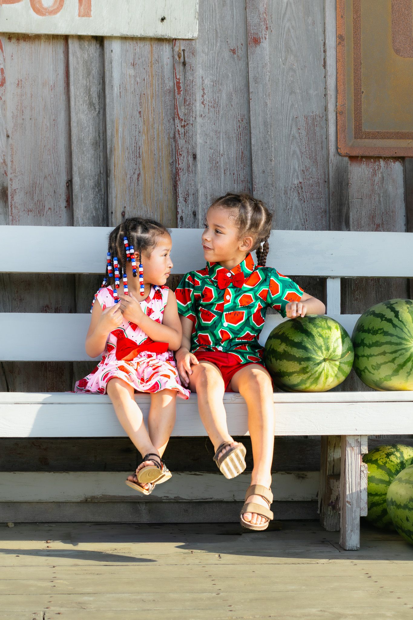 Two young girls in colorful outfits sit on bench chatting beside watermelons at farm stand.
