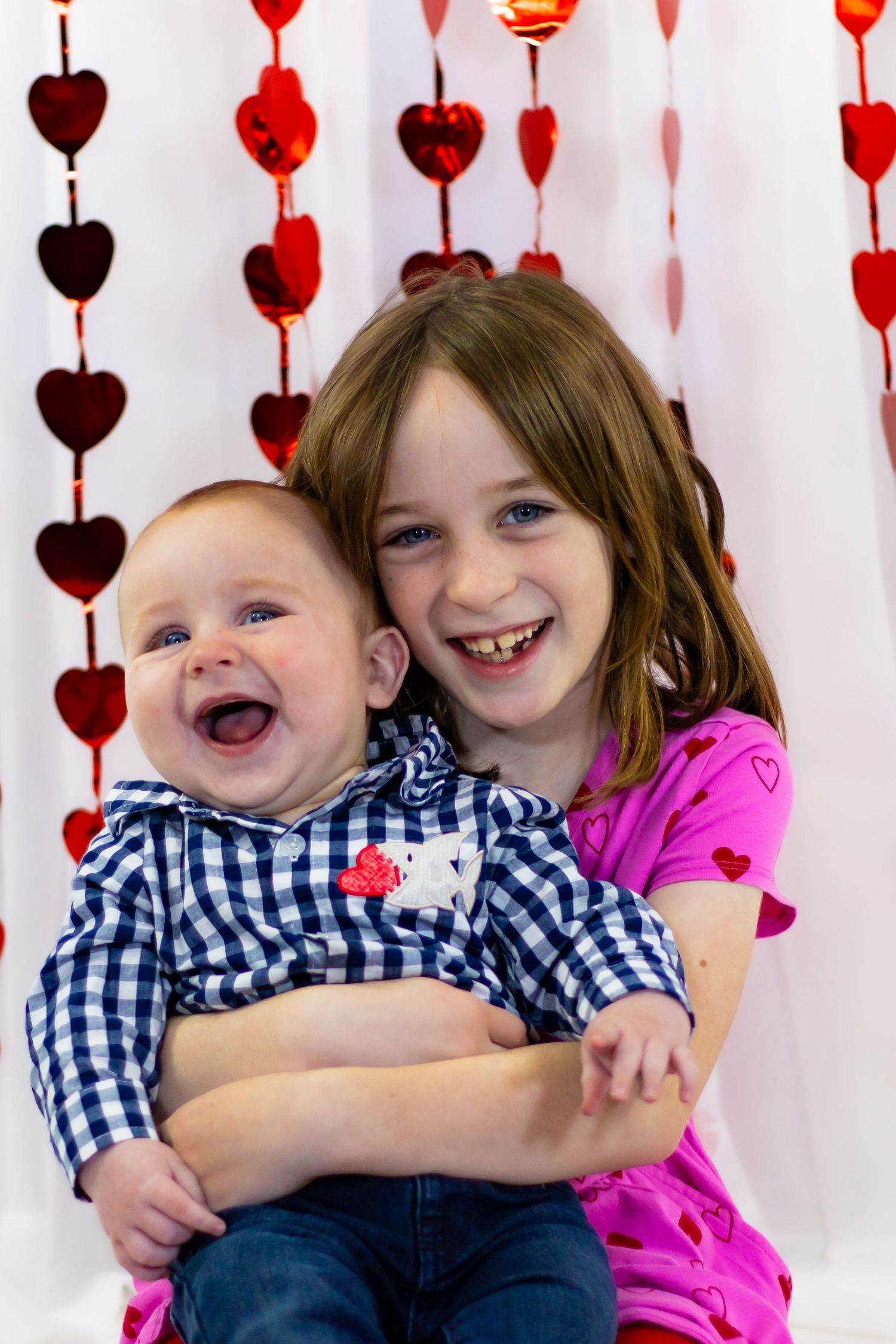 A smiling girl in pink holds a laughing baby in a plaid shirt against a Valentine's Day heart backdrop.