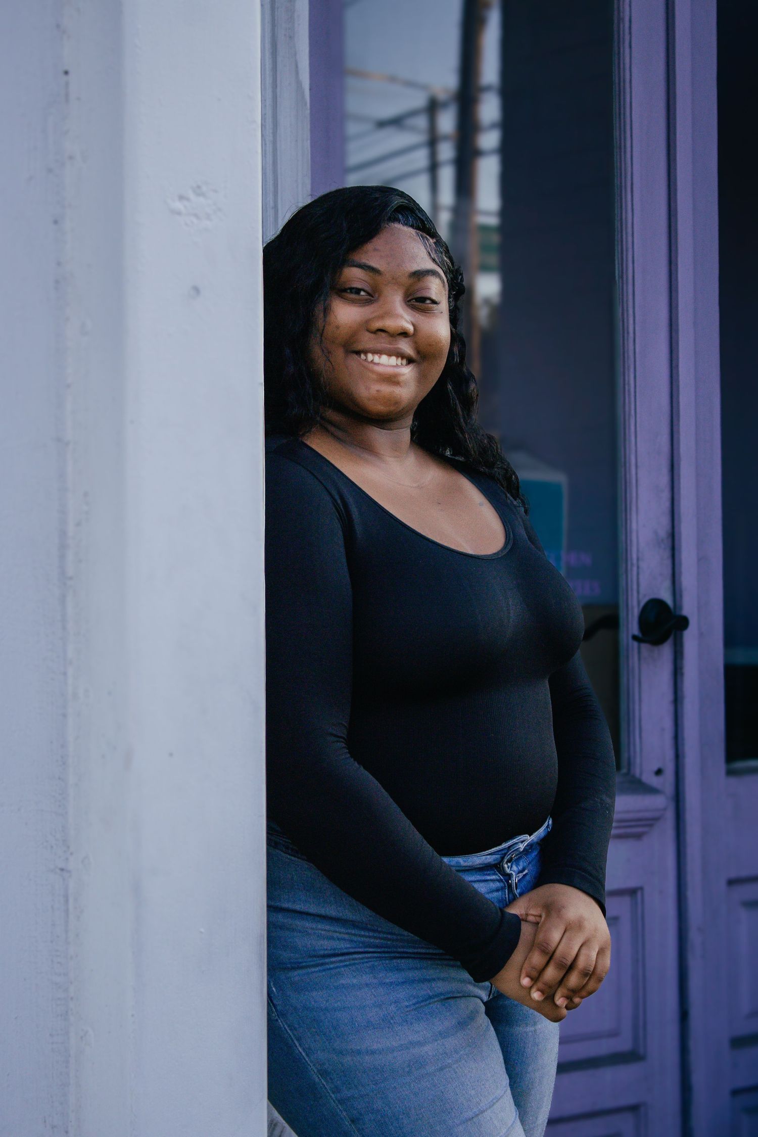 Smiling young Black woman in black long-sleeve top and jeans leaning against wall by purple door.
