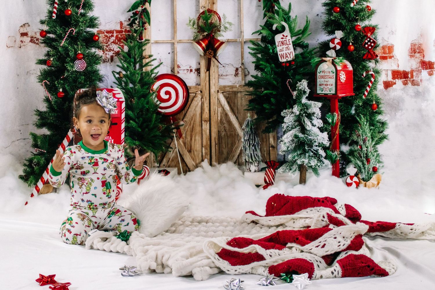 Toddler girl in Christmas pajamas smiling in festive holiday photo studio with trees and decorations.