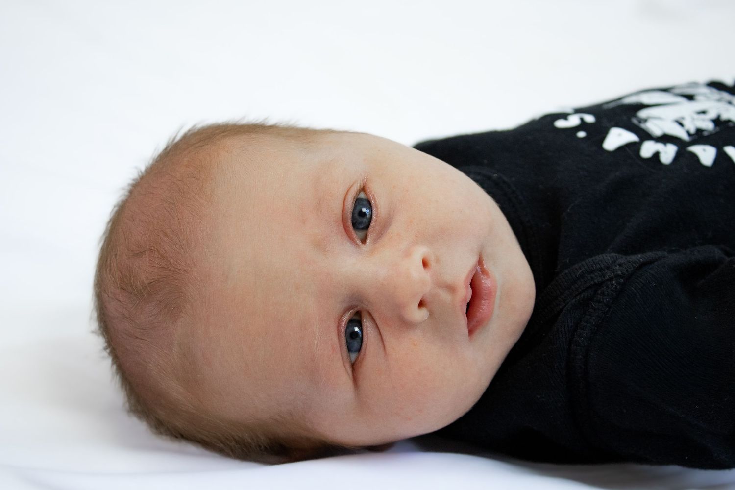 Newborn baby boy in black onesie lying down with bright blue eyes open.