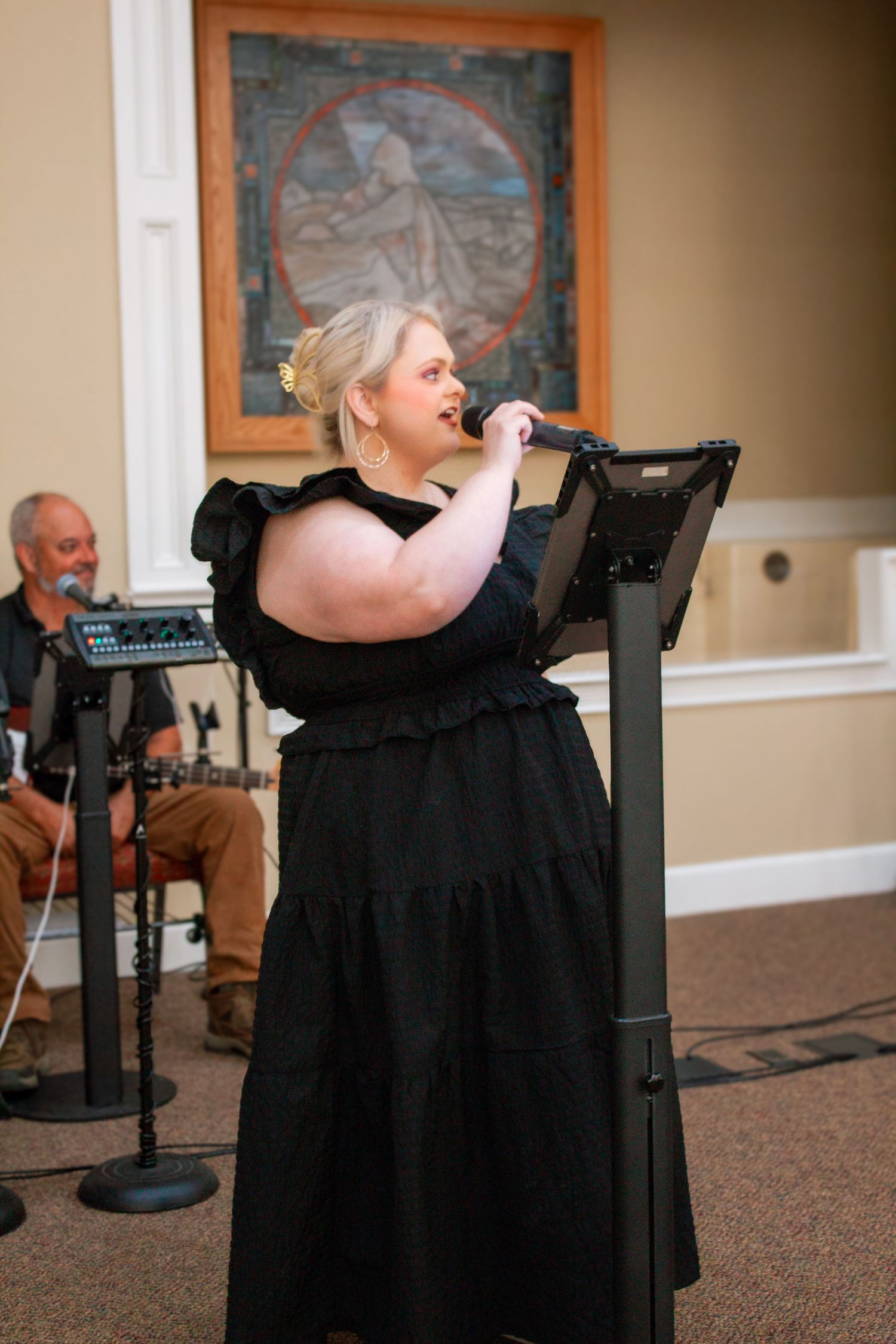 A plus-size blonde woman in a black dress sings into a microphone at a church worship service.