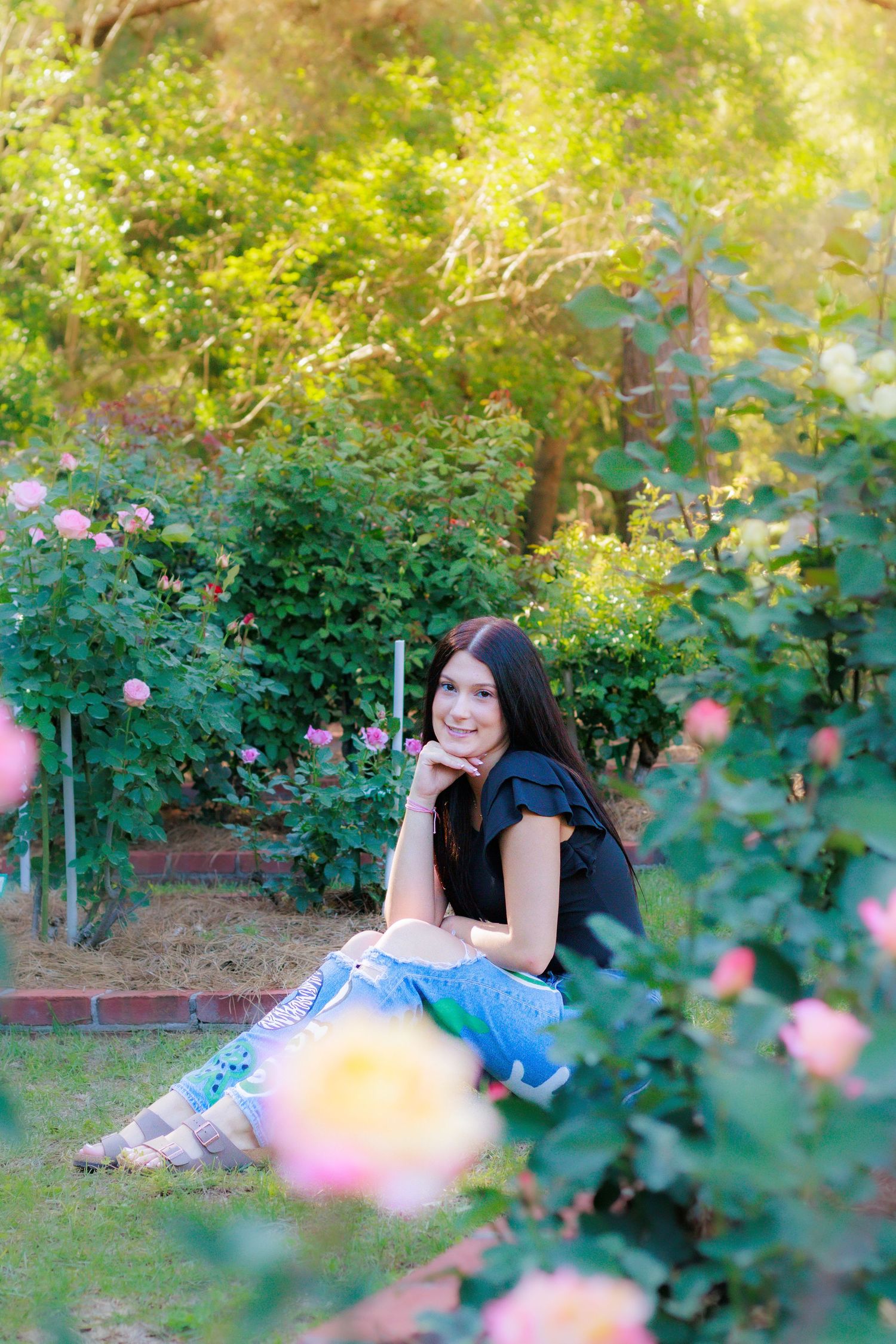 Young woman with long dark hair sits smiling among pink roses in a lush green garden wearing a black top and jeans.