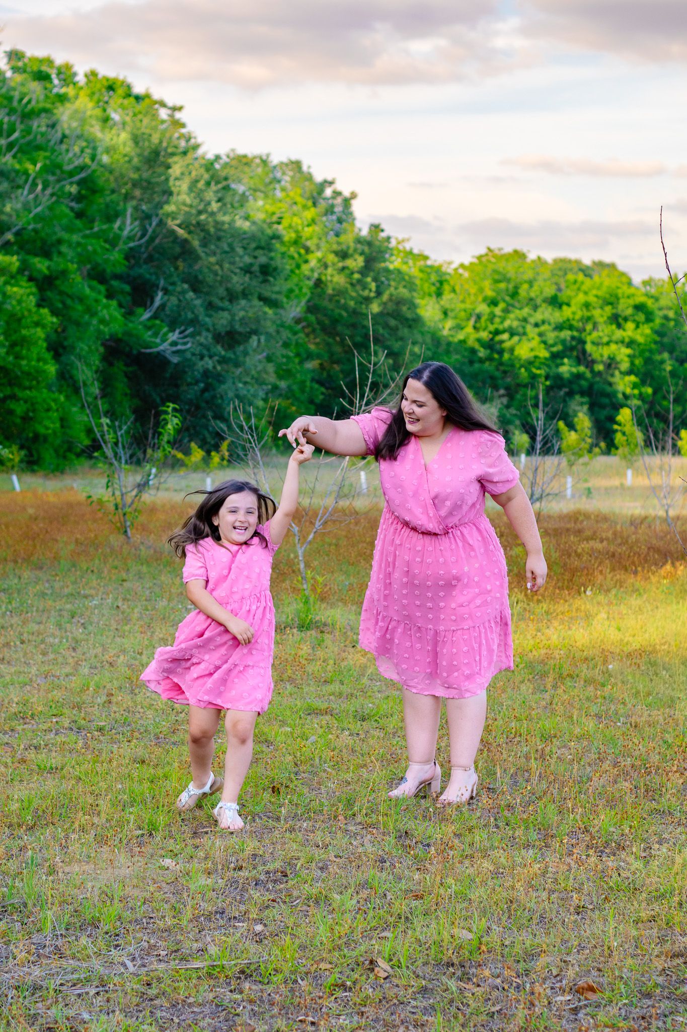 Mother and daughter in matching pink dresses dancing in a green field at sunset.