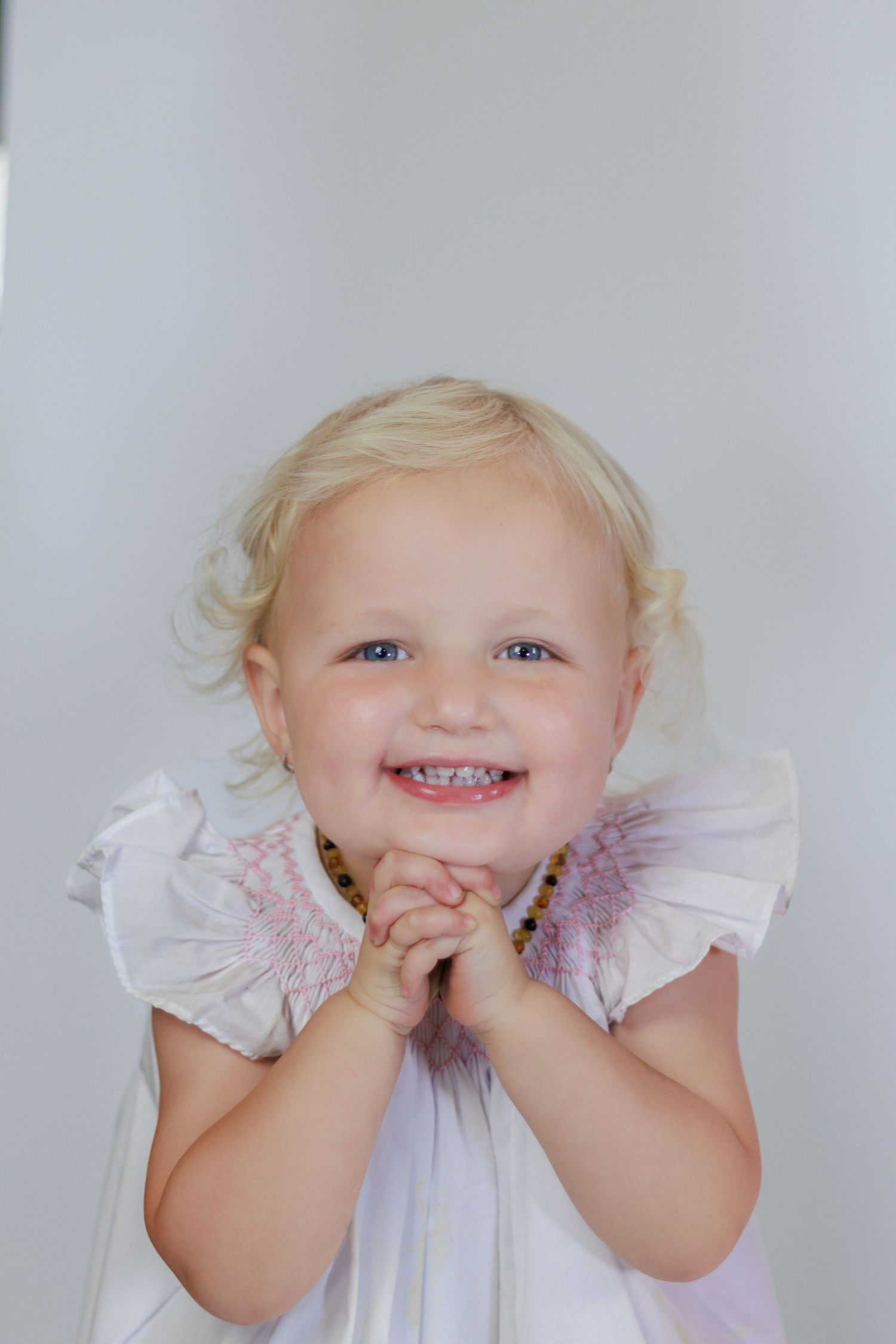 Adorable blonde toddler girl in white dress with purple flowers, smiling with hands clasped together.