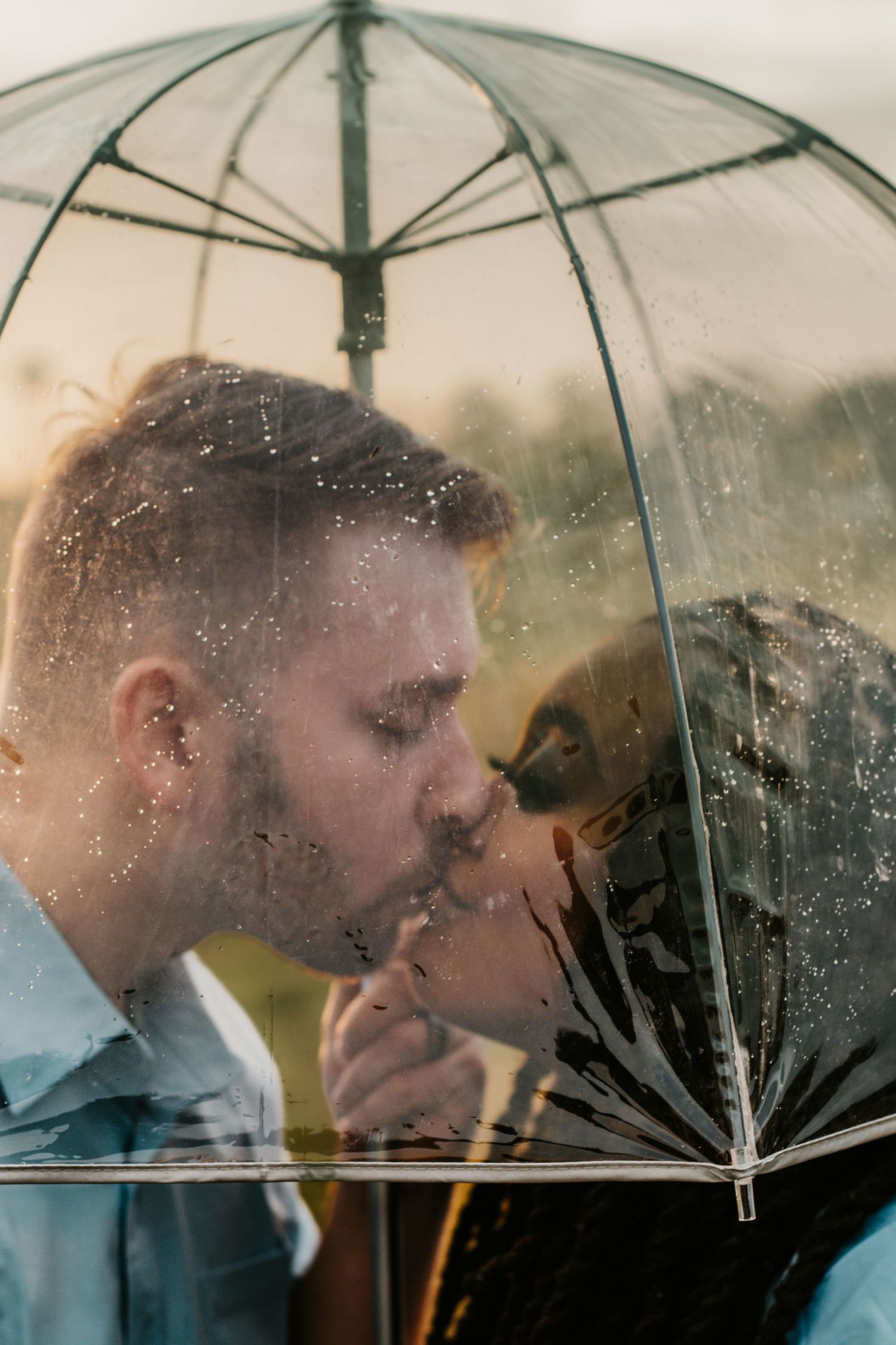 Couple sharing a romantic kiss under a clear transparent umbrella in the rain at sunset.