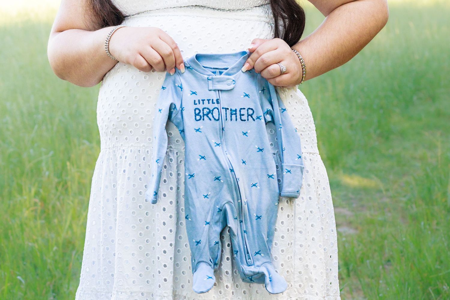Pregnant woman in white dress holding a blue Little Brother baby onesie over her bump outdoors.