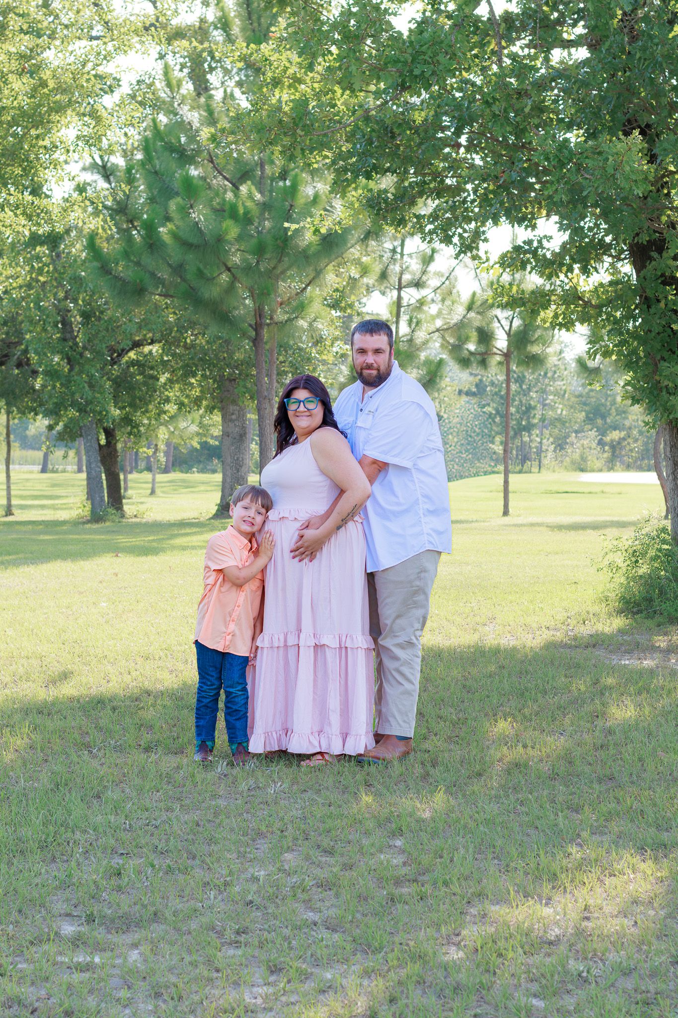 A pregnant woman in a pink dress poses with her husband and young son in a lush green park setting.