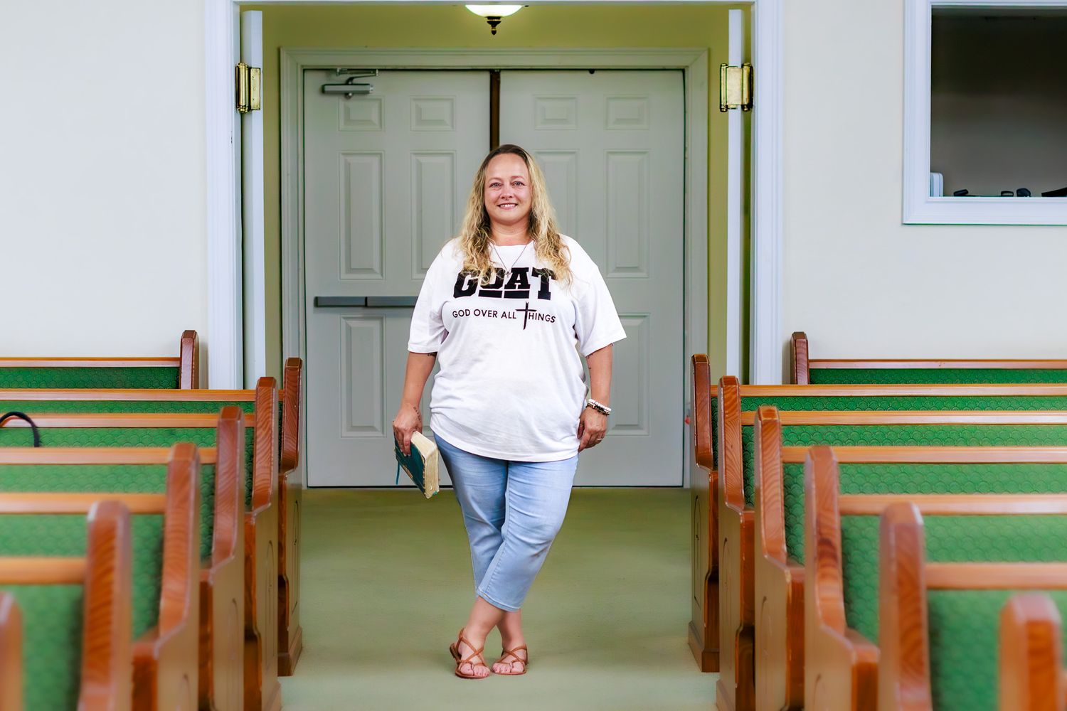 Woman in GOAT t-shirt and jeans poses confidently between wooden church pews in a small chapel.