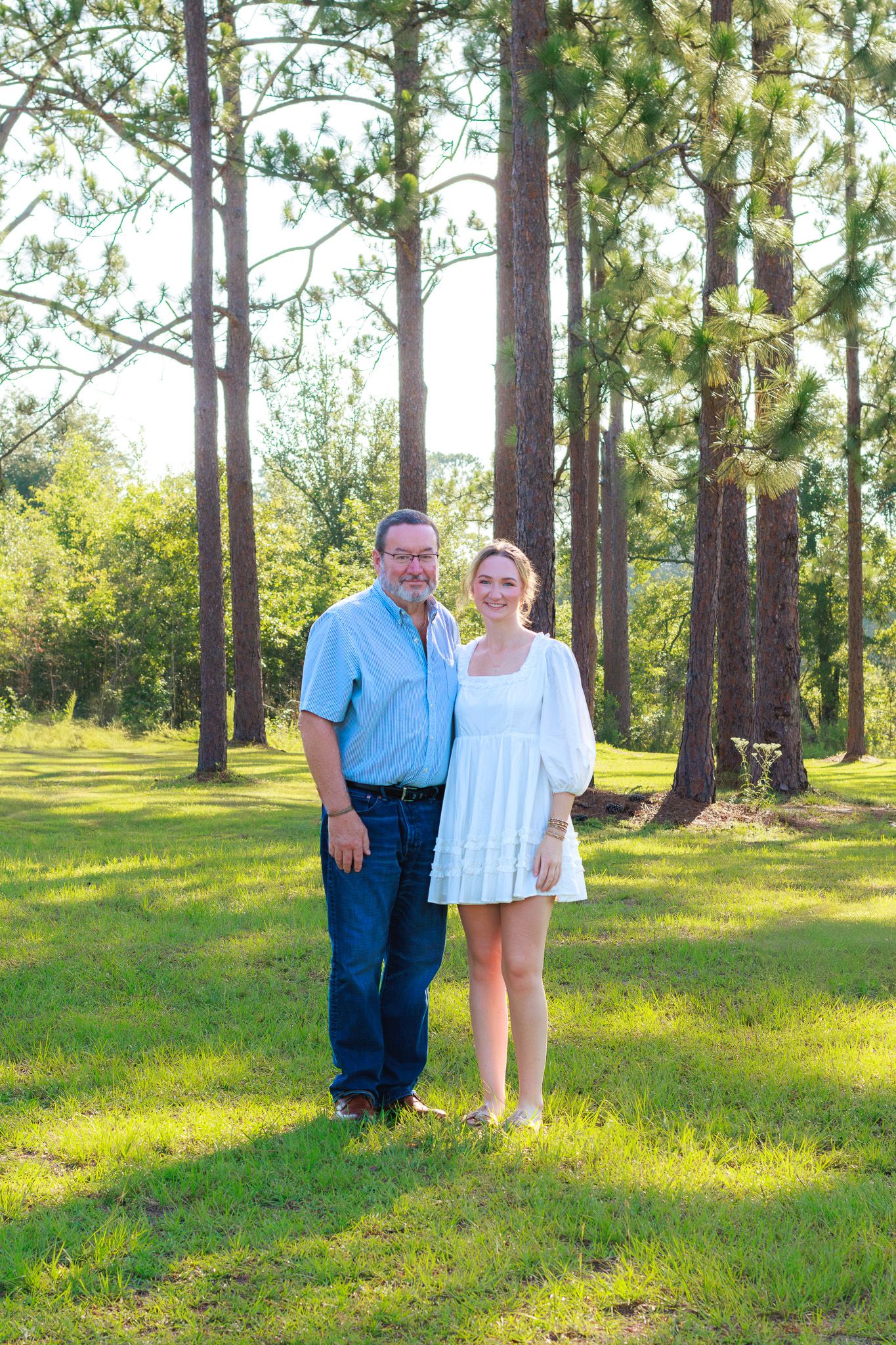 Smiling couple stands together on green lawn surrounded by tall pine trees in natural outdoor setting.