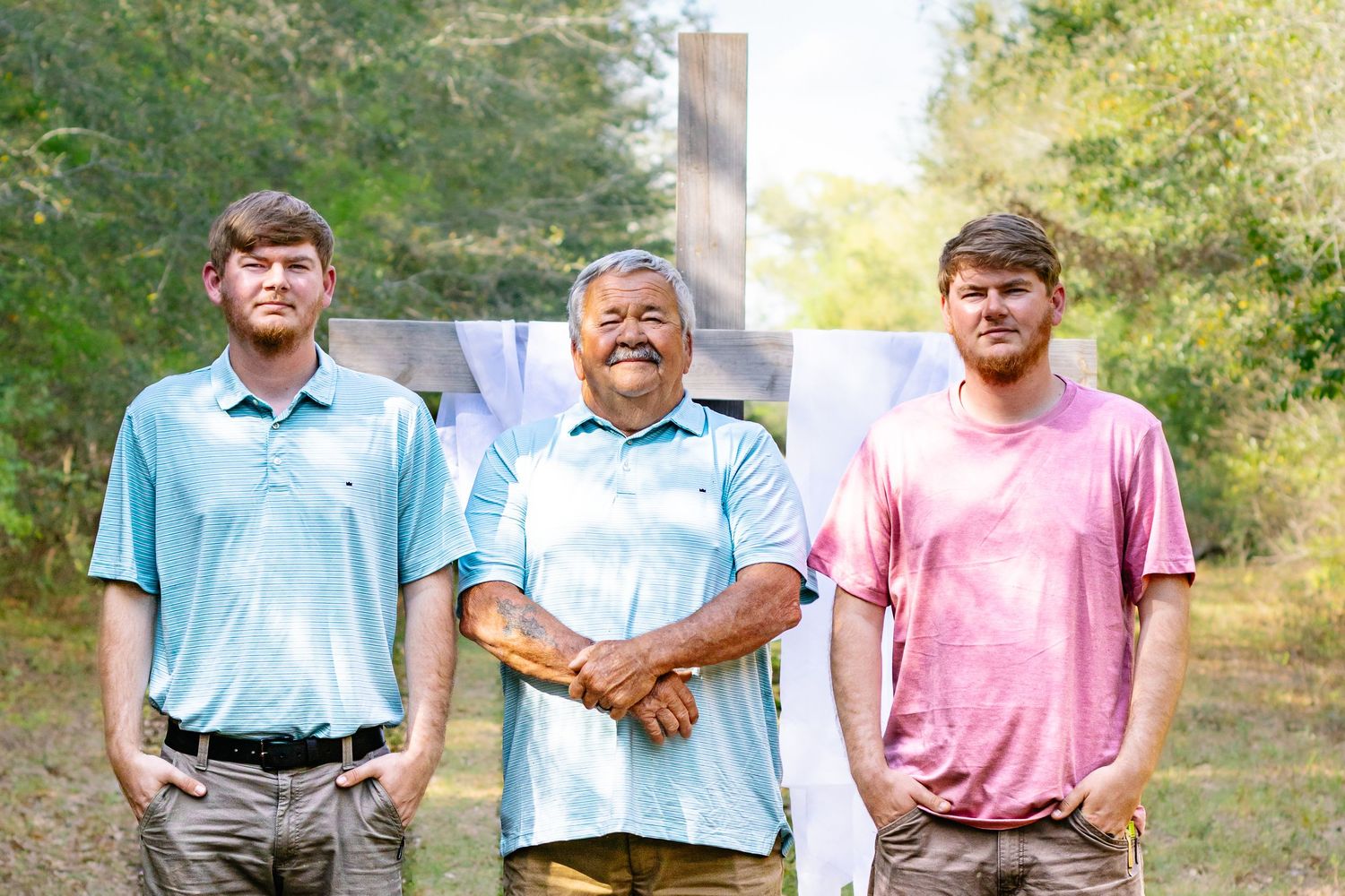 Three men standing together outdoors in front of a wooden cross on a sunny day.
