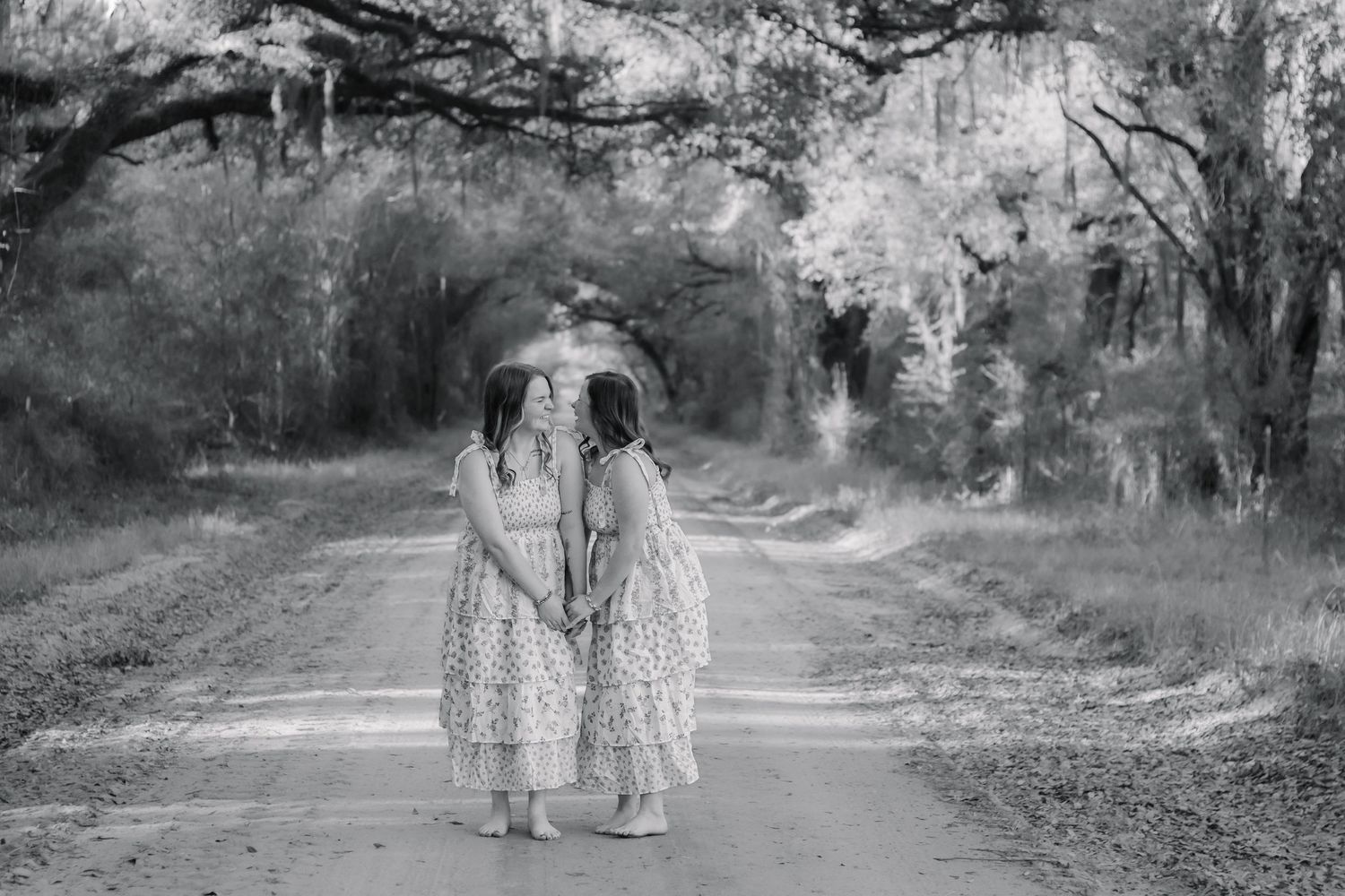 Black and white photo of two girls in floral dresses walking hand in hand on a dirt road.