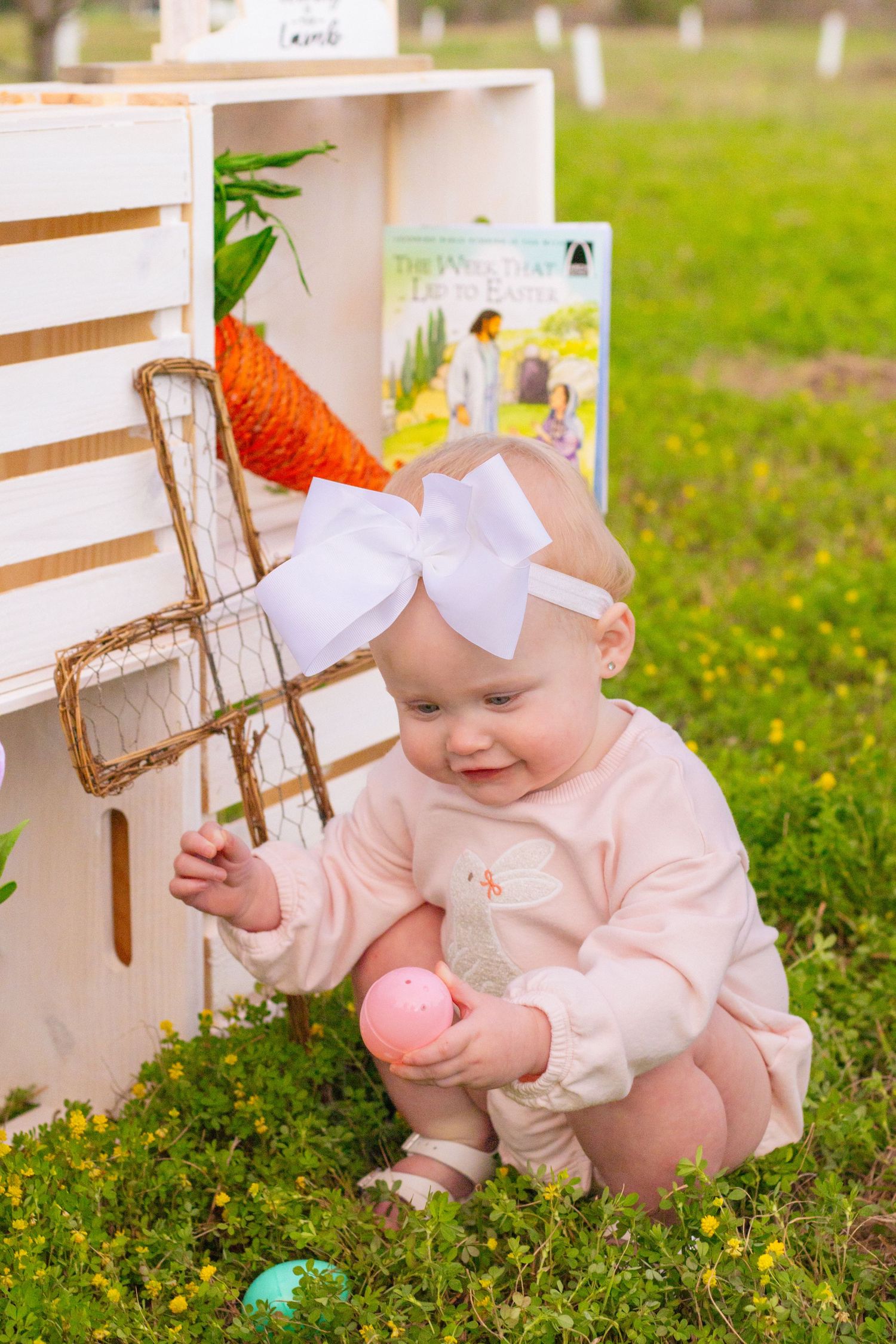 Smiling baby with white bow headband holds pink Easter egg near white wooden crate.