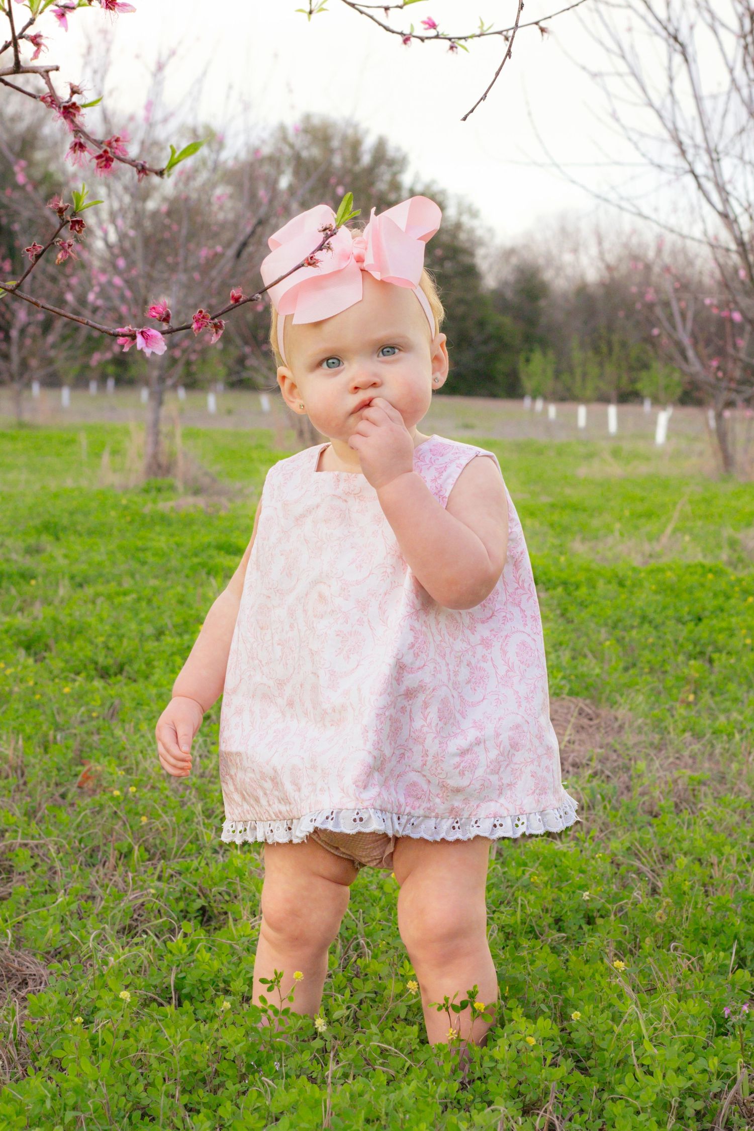 Cute baby girl in pink dress with bow headband standing near blooming cherry blossom branch in garden.